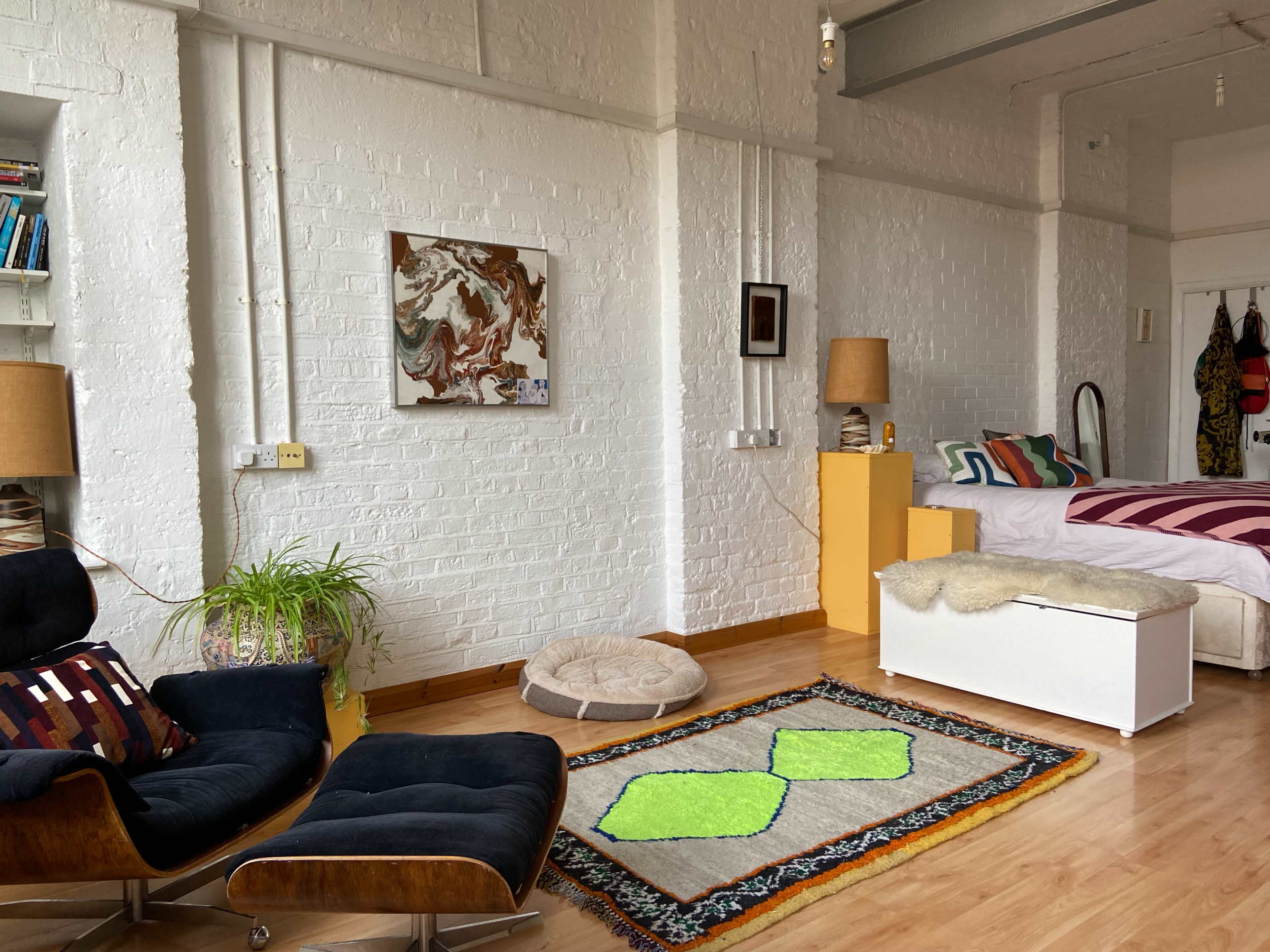 A bright, airy studio apartment features a bed, a black armchair, a colorful rug with green accents, and artwork on the white brick walls.