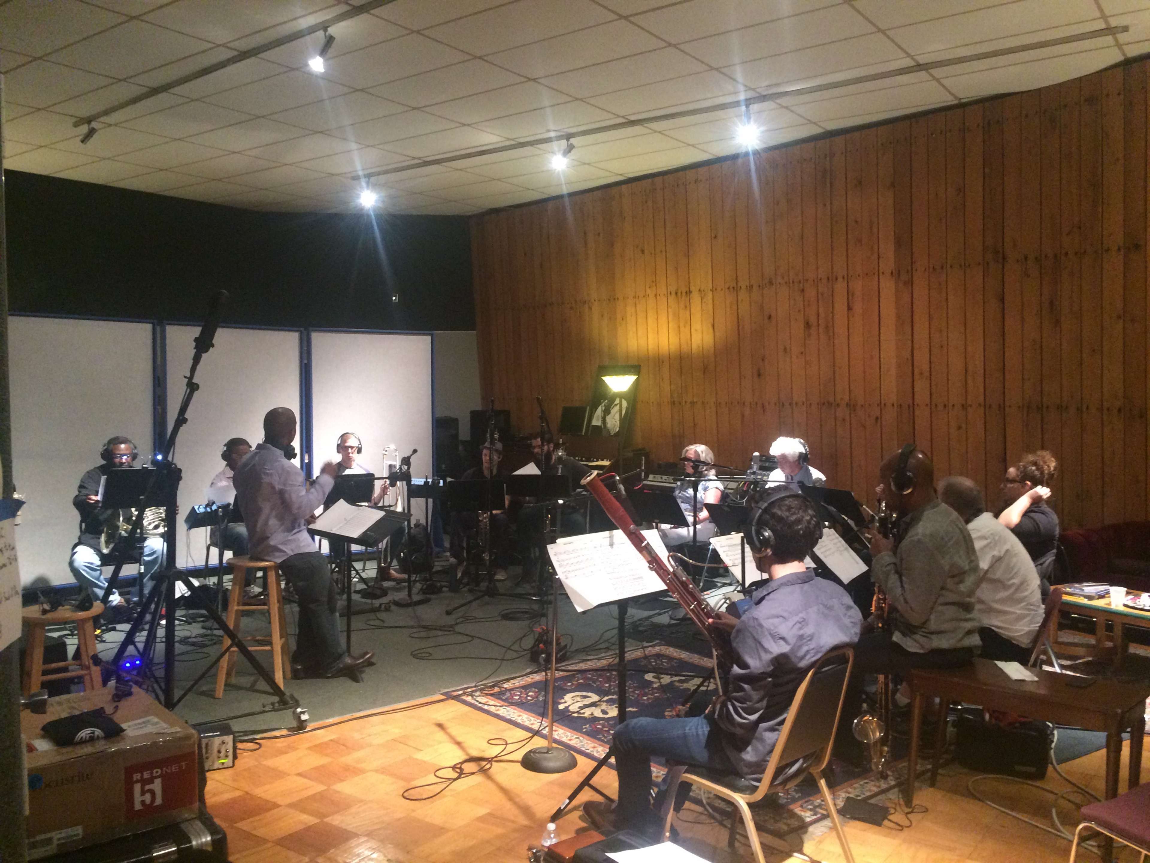 A group of musicians sits in a recording studio, arranged in a semicircle around a conductor who is leading the session.