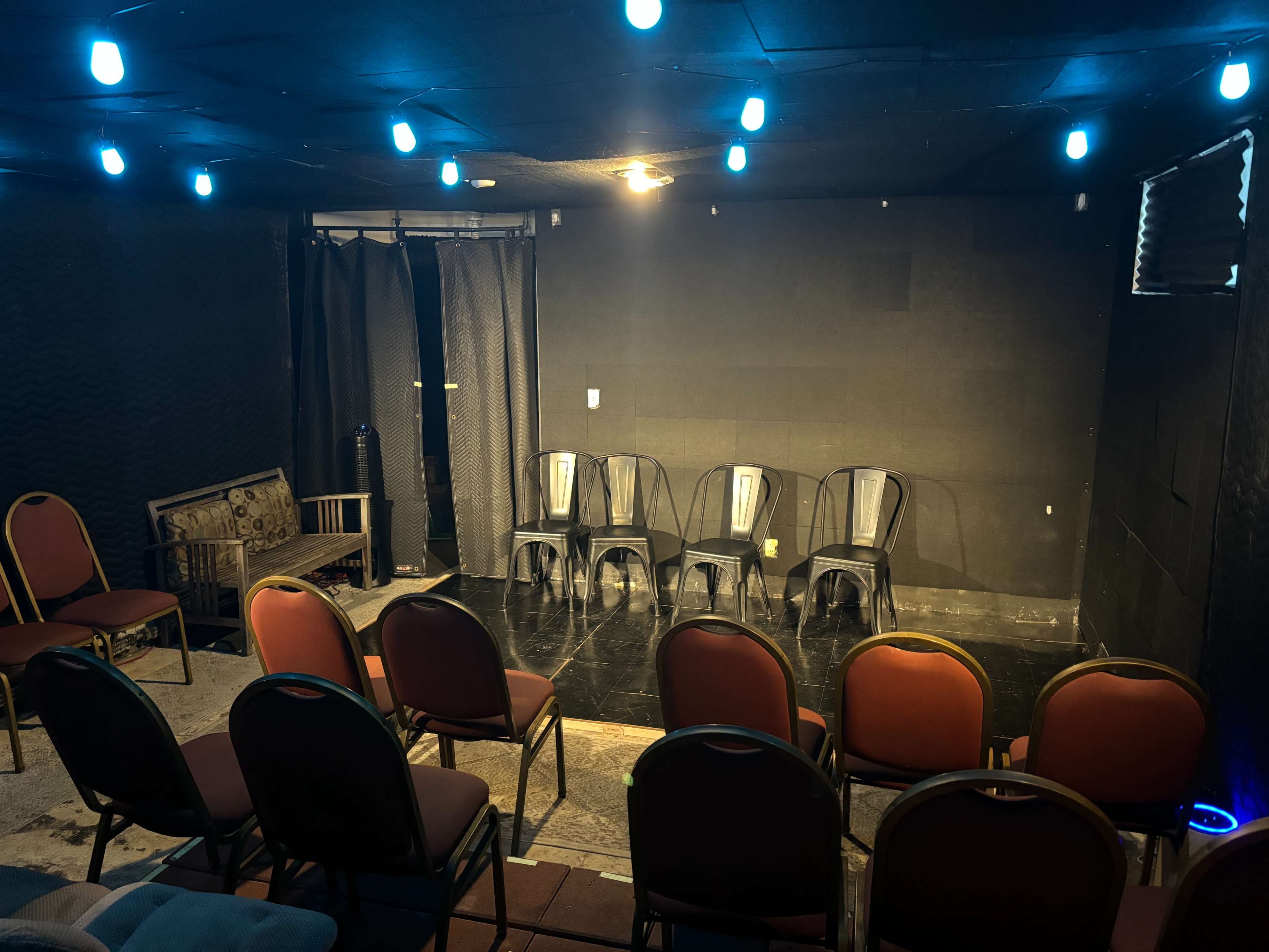 A small performance space with chairs arranged in a semi-circle facing a stage area, illuminated by blue lights.