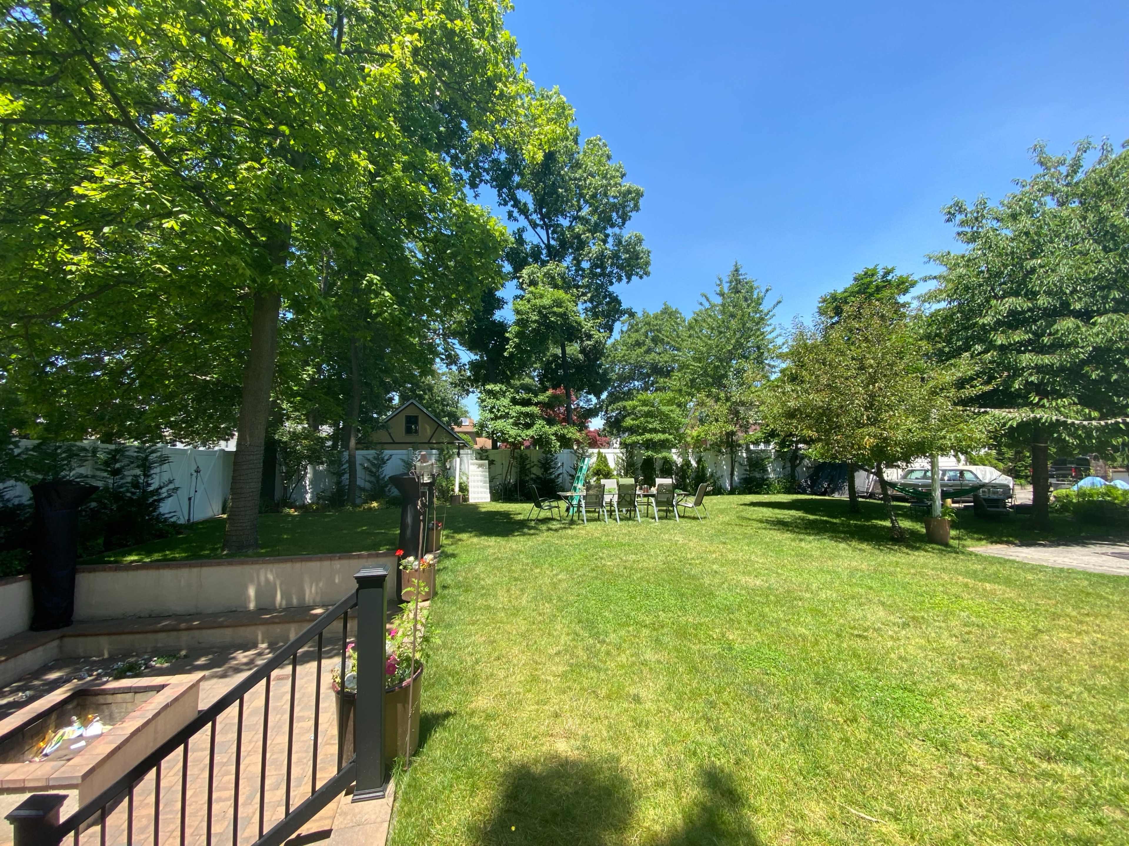 A spacious backyard features green grass, several trees, and a set of chairs arranged under the sunlight.