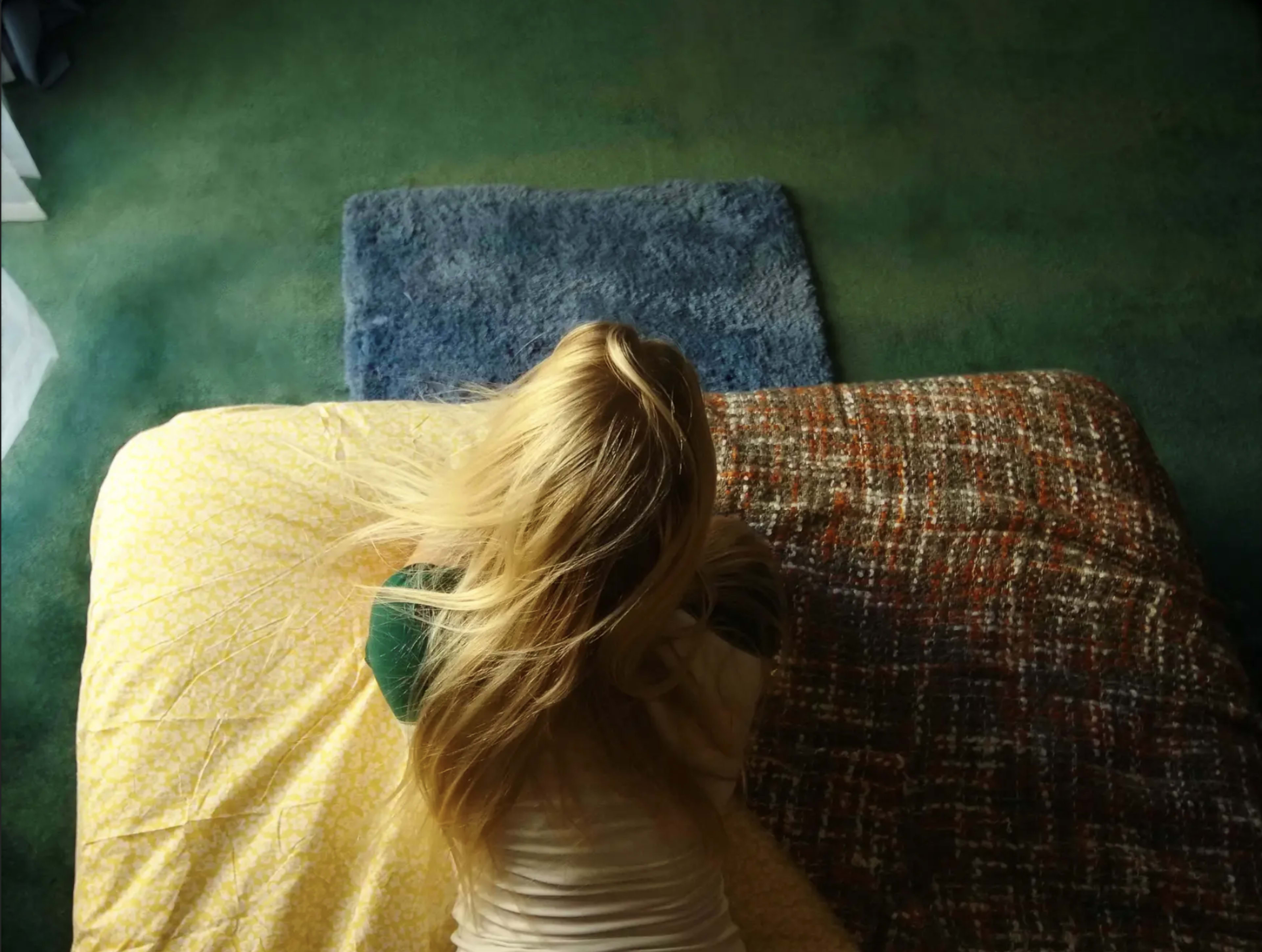 A person with long hair is sitting on a bed, facing away, with a blanket partially covering them.