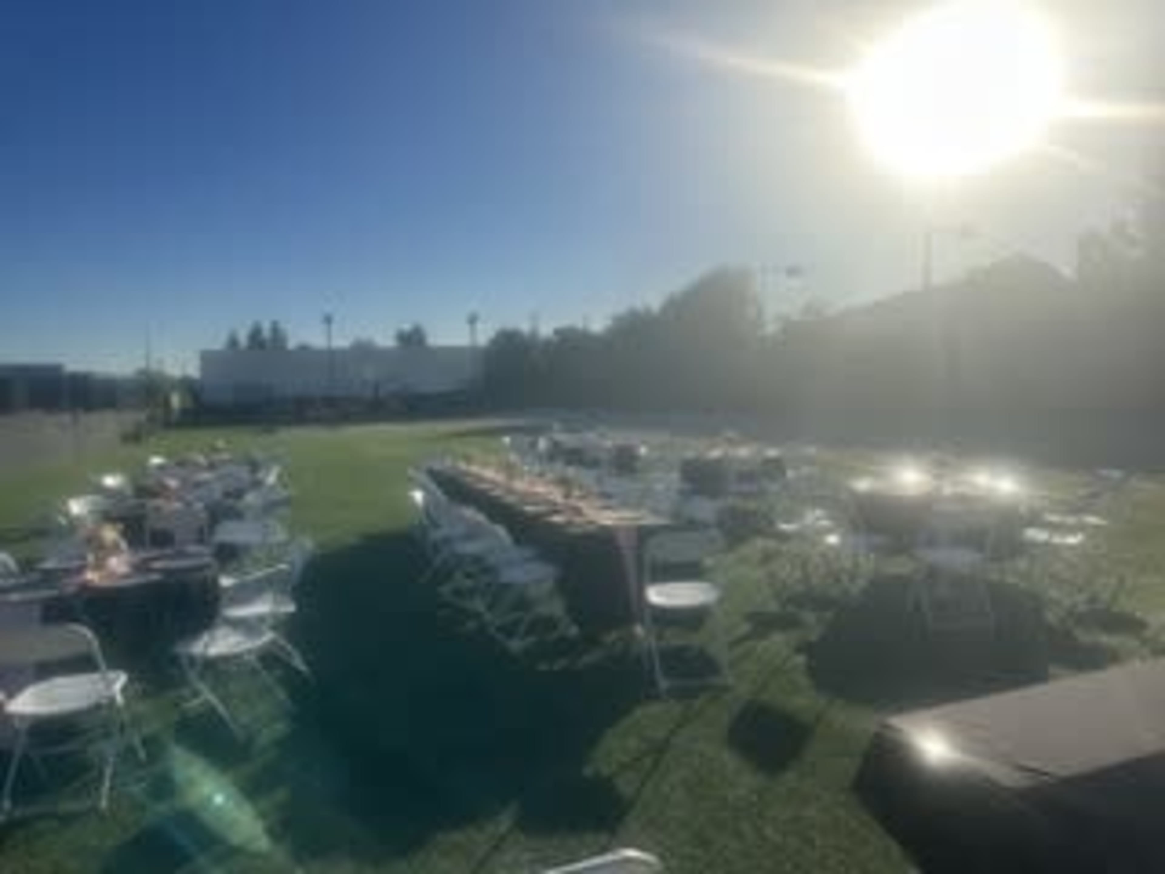 The image shows an outdoor event setup with several round tables and chairs arranged on a grassy area under clear blue skies.