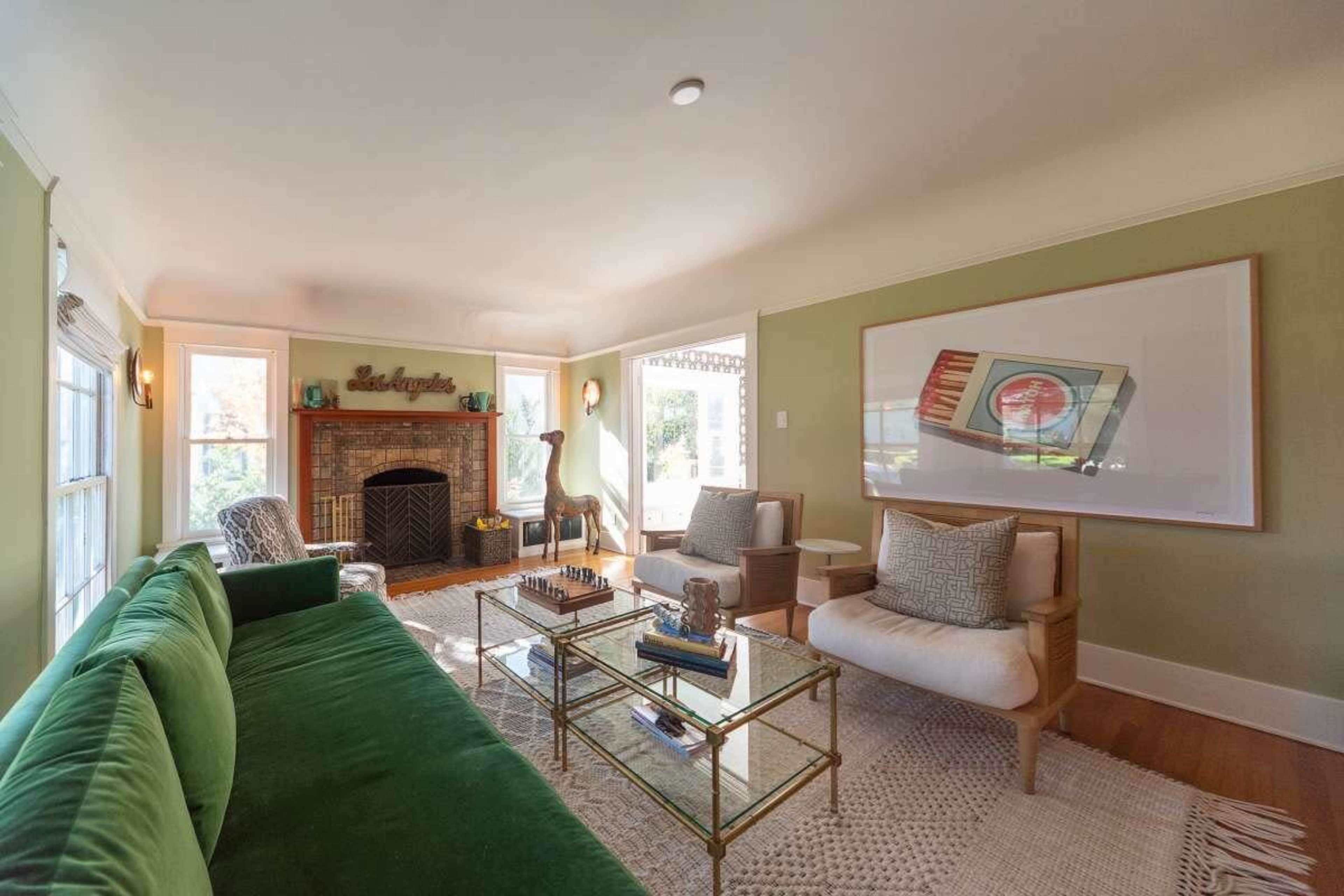 The image shows a living room with a green sofa, a coffee table, and a fireplace, surrounded by large windows that let in natural light.