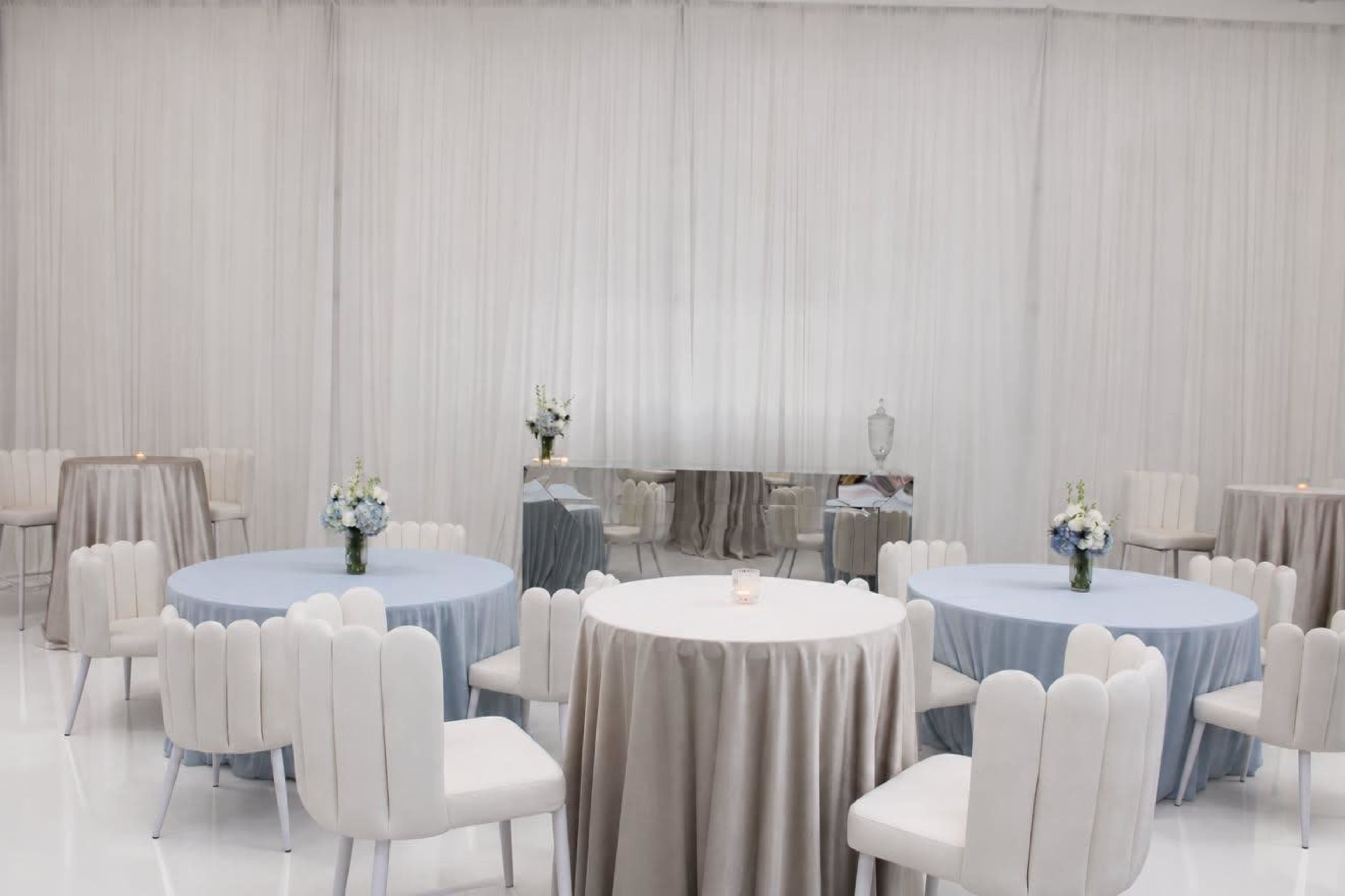 The image shows a elegantly decorated room with round tables covered in light blue and beige tablecloths, surrounded by chairs, and complemented by floral centerpieces.