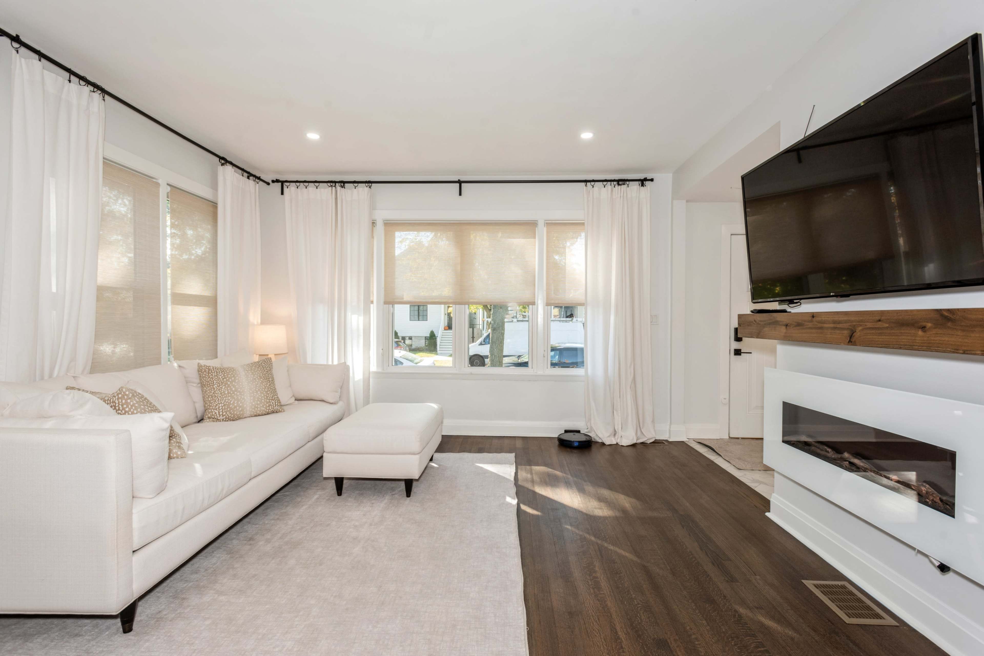 A modern living room with a white sectional sofa, a mounted television, and large windows covered with sheer curtains.