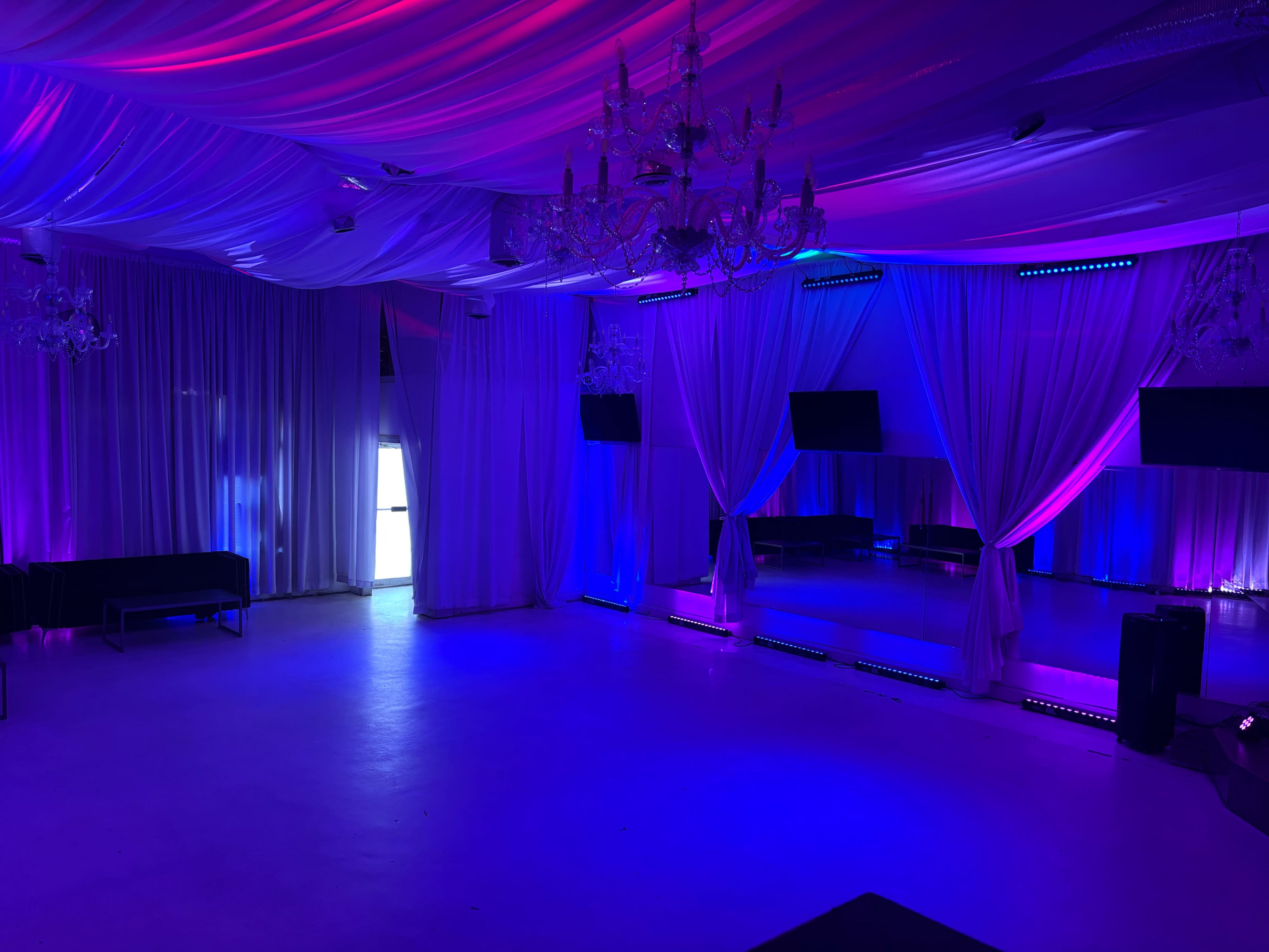 The image shows a spacious event room with draped fabric and colorful lighting, featuring a mirrored wall and chandeliers.