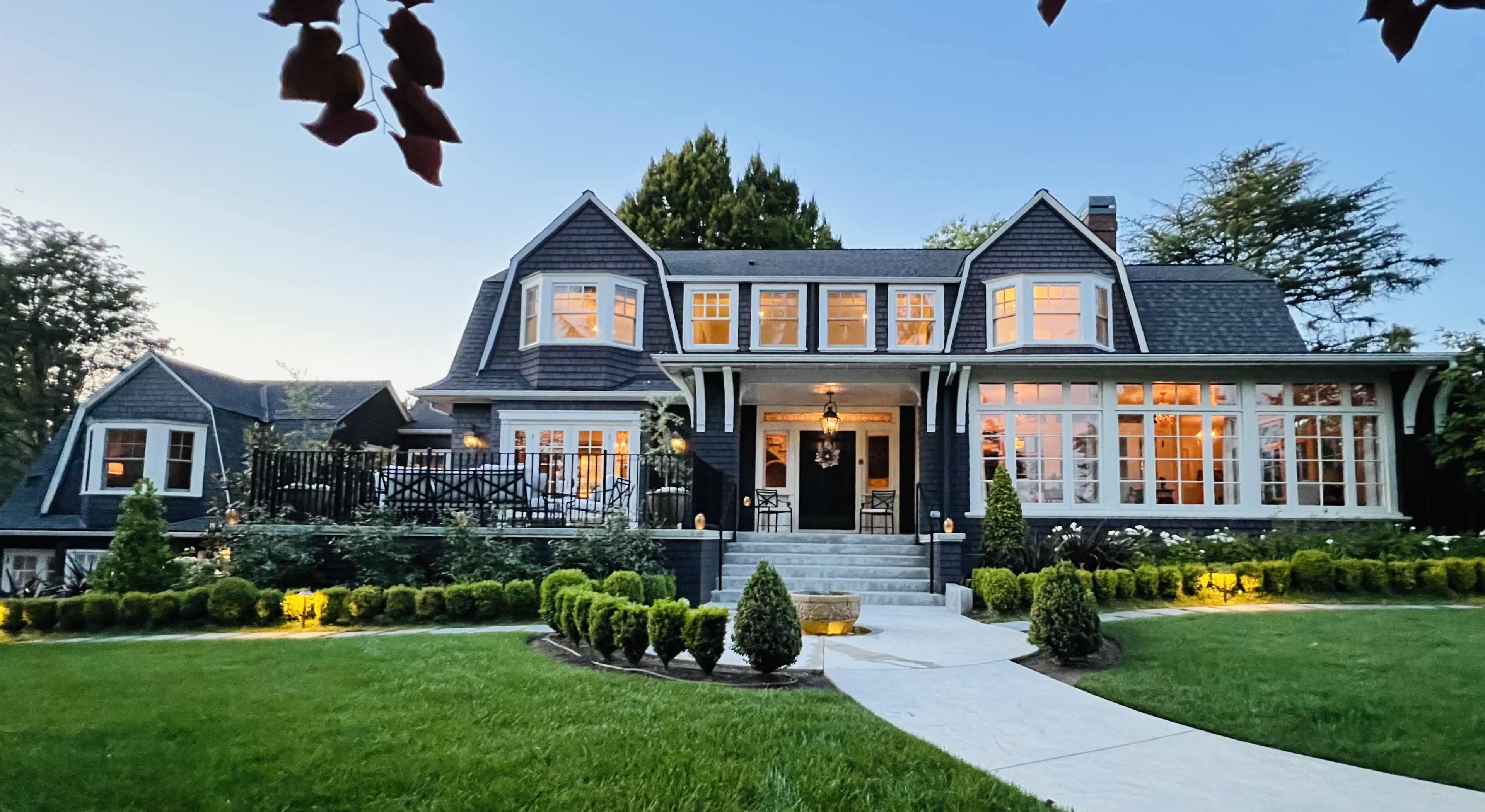 A two-story house with a dark exterior and multiple large windows is surrounded by neatly trimmed hedges and a well-kept lawn during twilight.