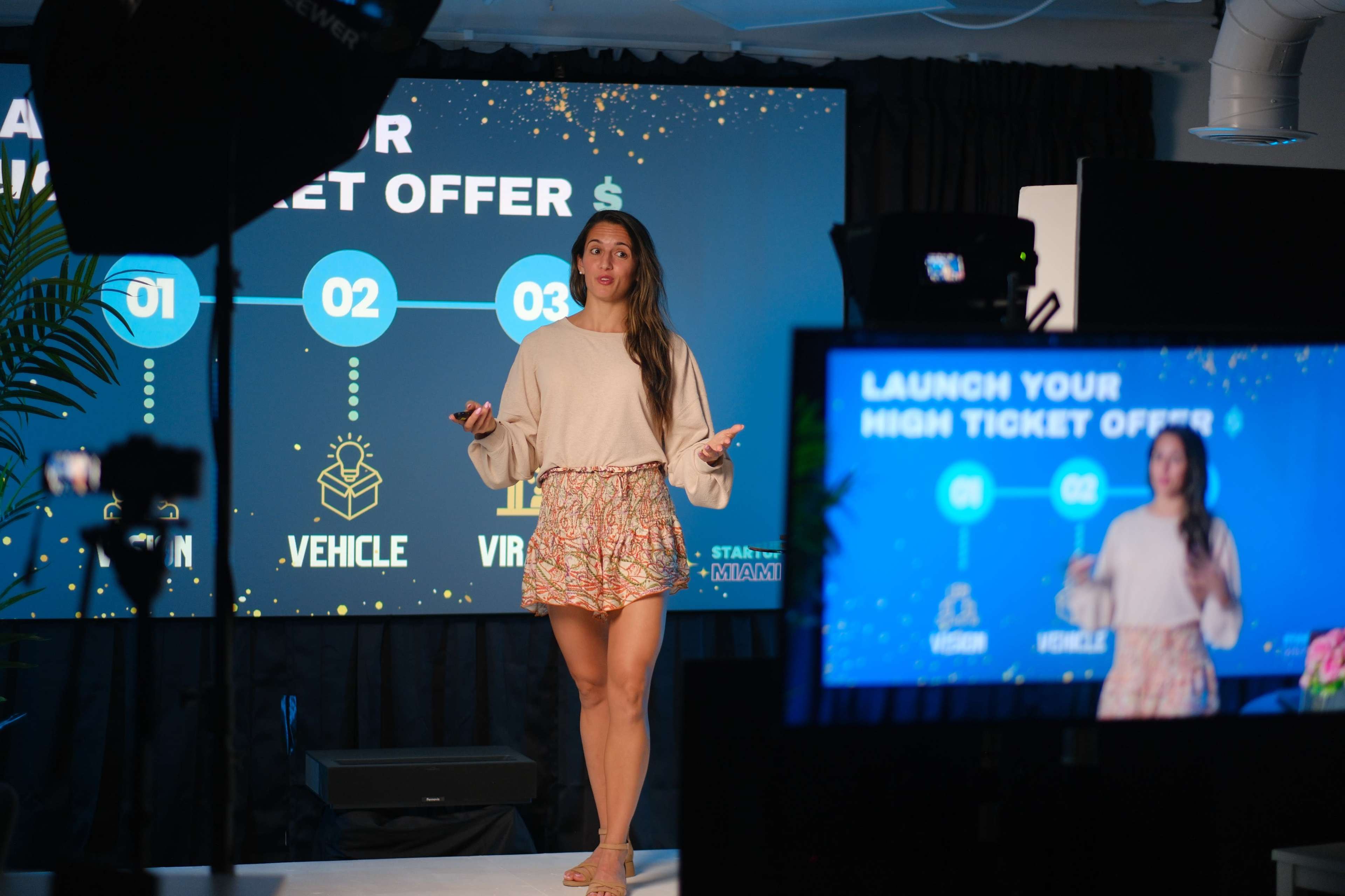 A woman stands on a stage in front of a large screen displaying a presentation about launching high ticket offers.