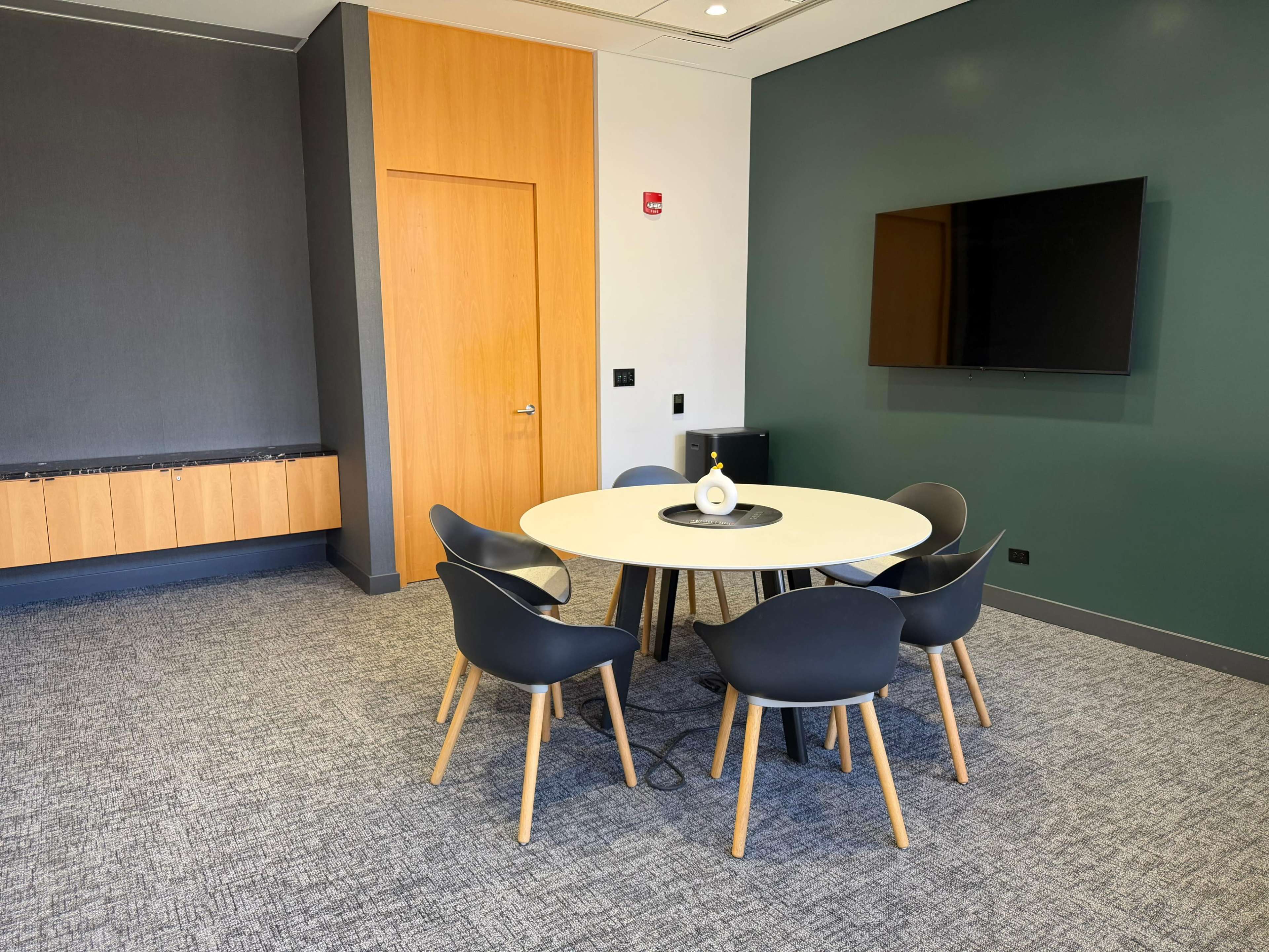 The image shows a modern meeting room with a round table and four black chairs, a wall-mounted television, and wooden accents.
