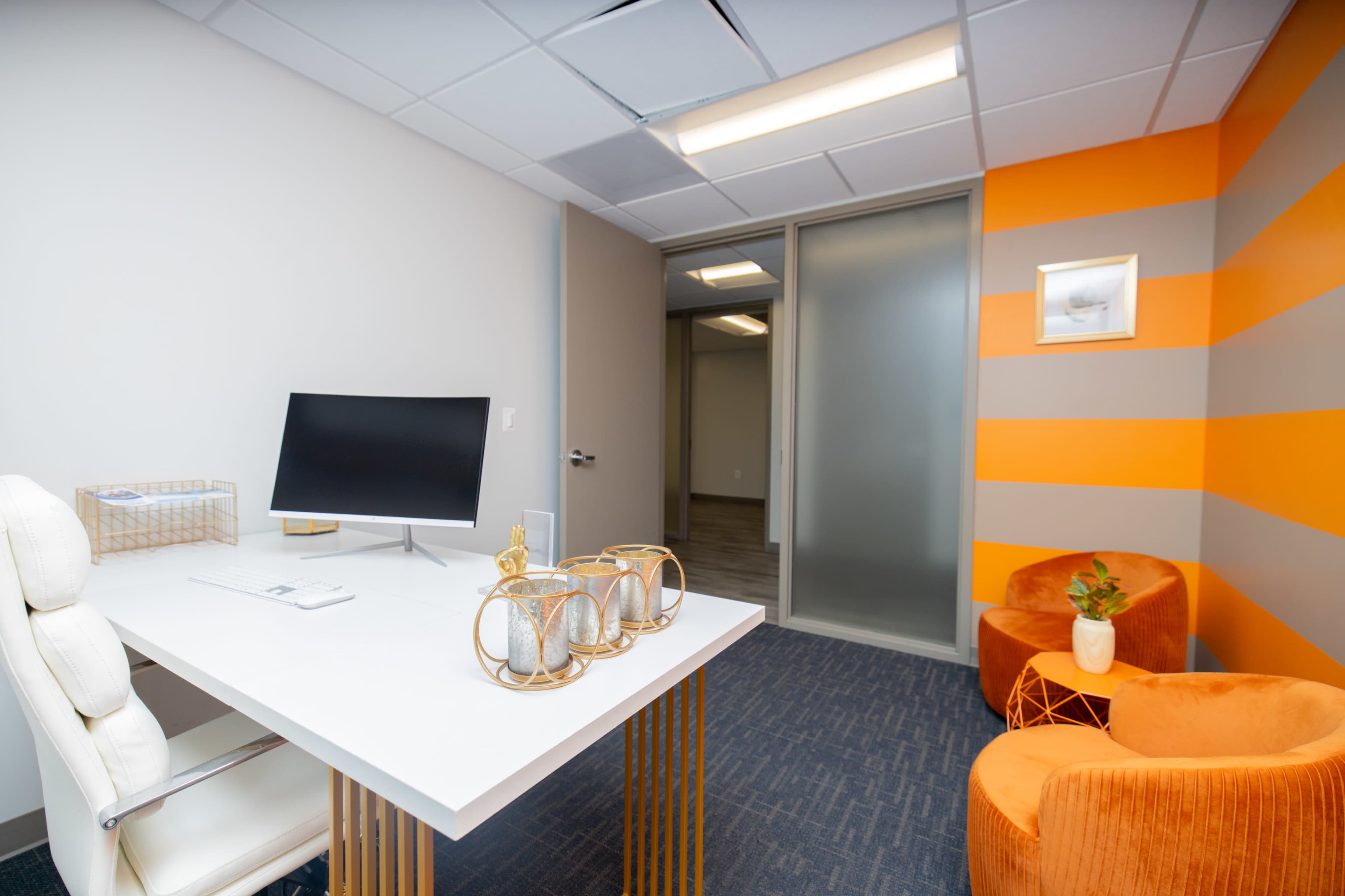 A modern office space features a desk with a computer, an armchair, and an orange accent wall with gray stripes.