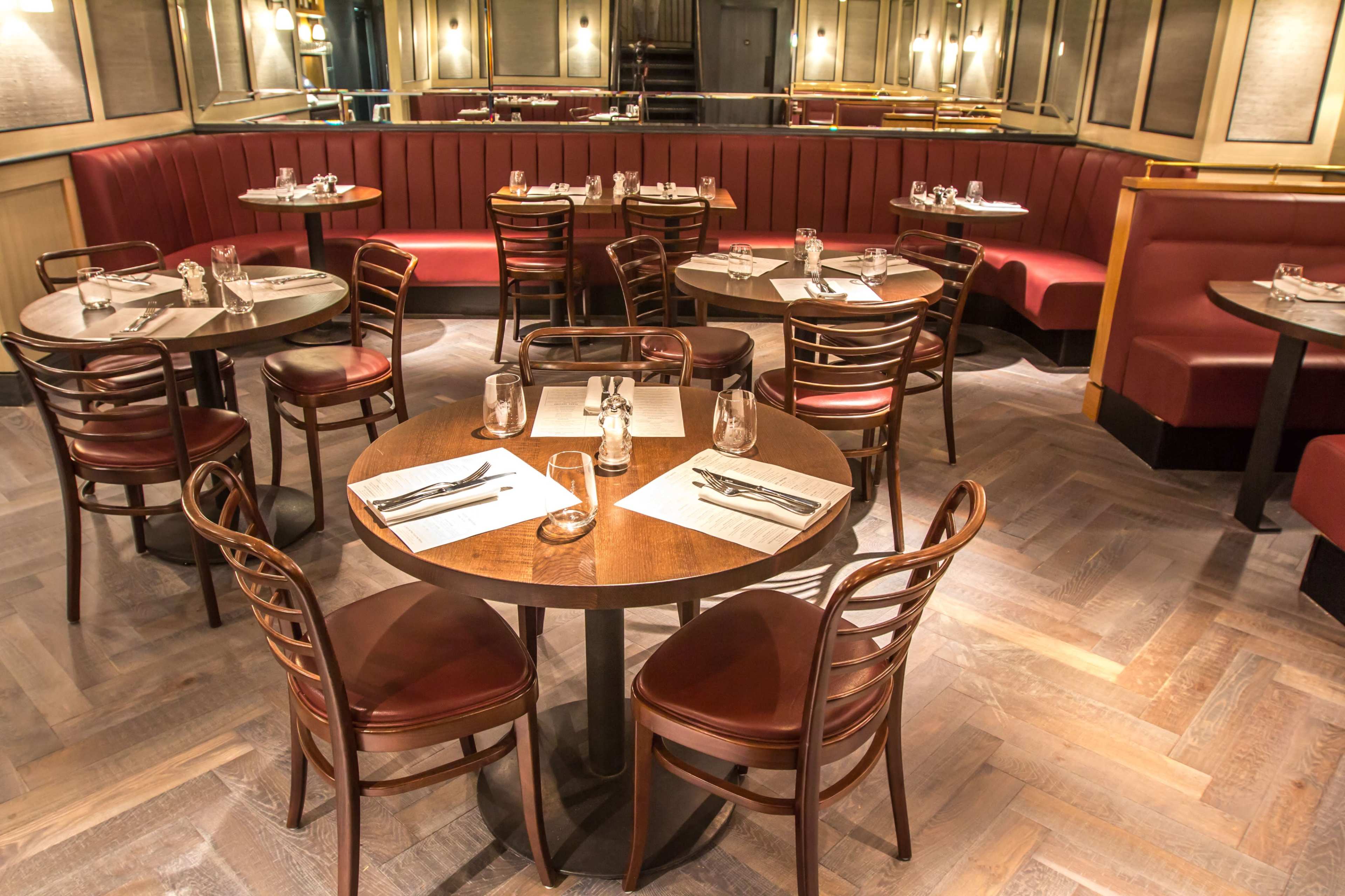The image shows a restaurant interior with several round wooden tables, red upholstered booths, and a herringbone-patterned wooden floor.