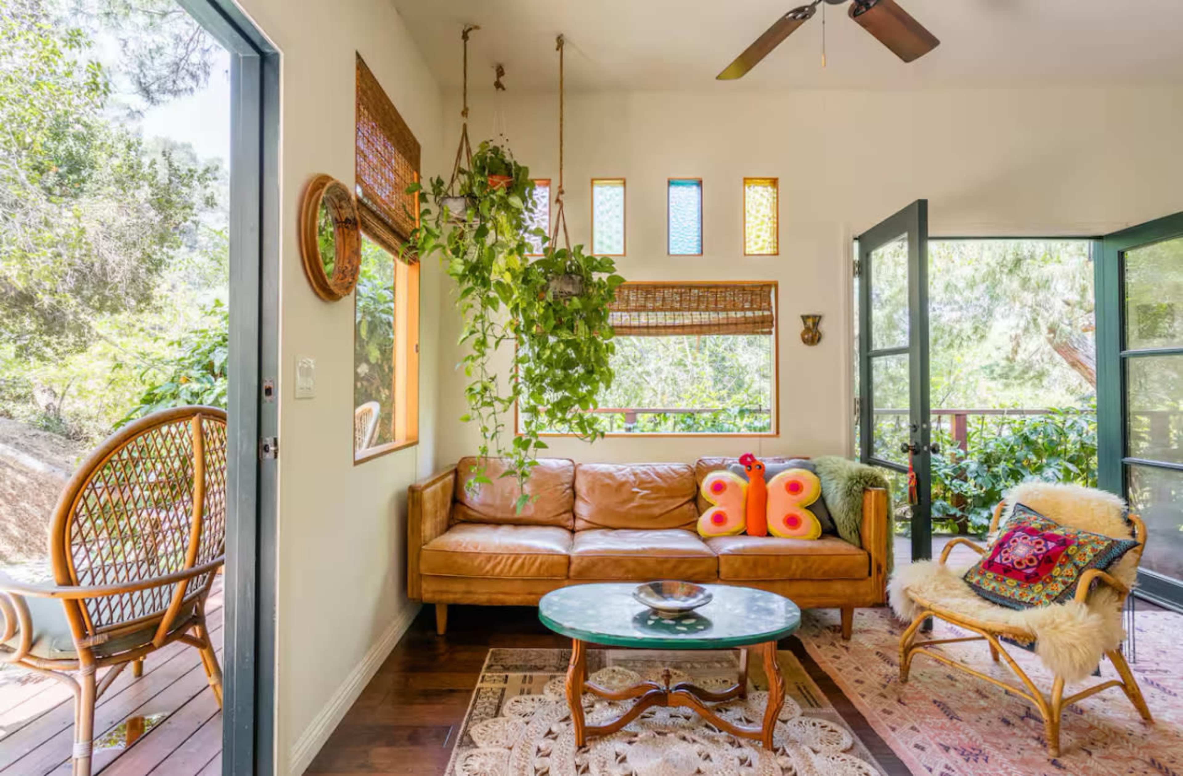 A cozy living room features a brown leather couch, a glass coffee table, and decorative plants, with large windows allowing natural light to fill the space.