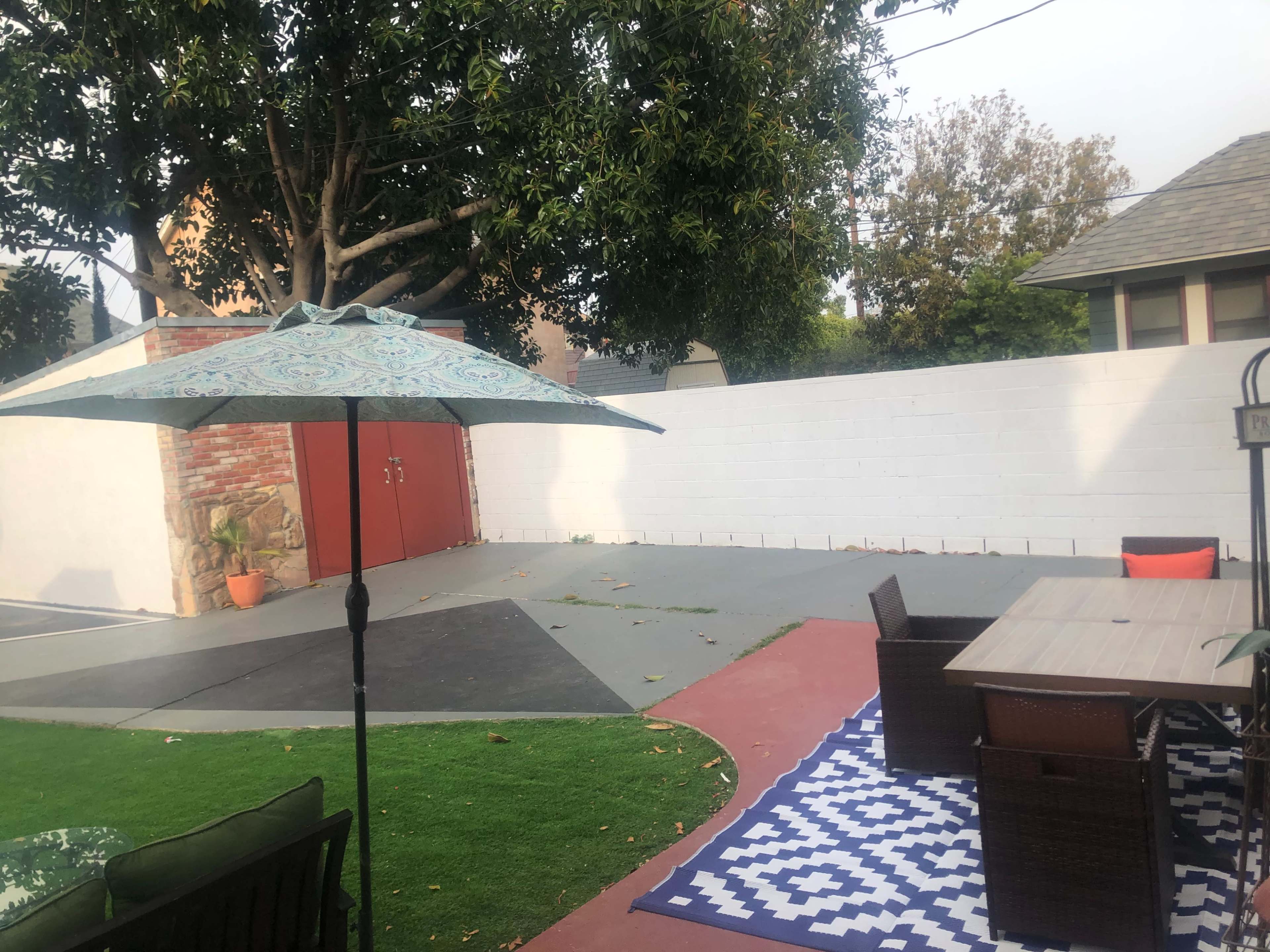 A patio area features a table and chairs, an umbrella, and a patterned rug on artificial grass, with a brick wall and trees in the background.