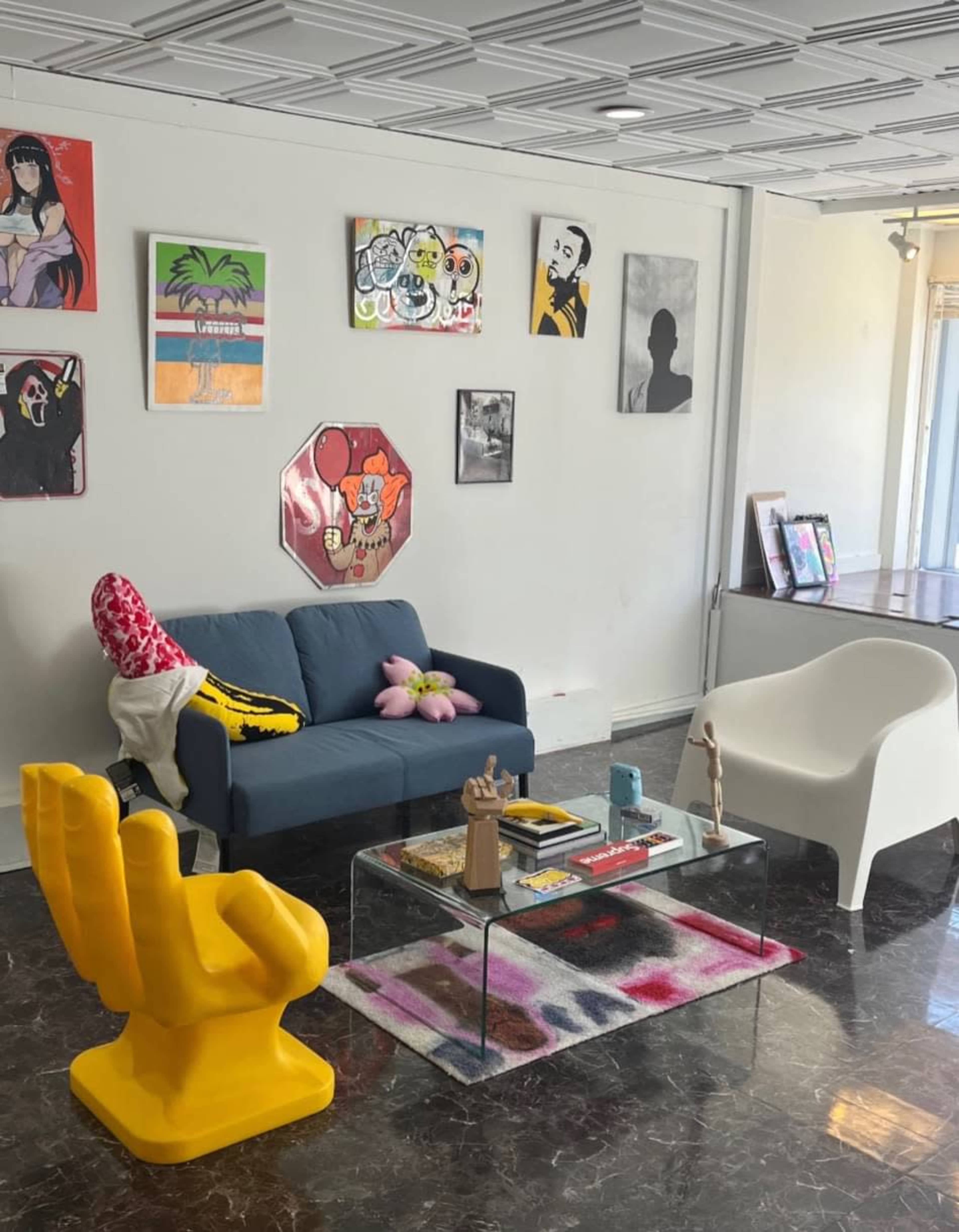 A modern living room with a blue couch, a yellow sculptural chair, a glass coffee table, and various artworks hanging on the walls.