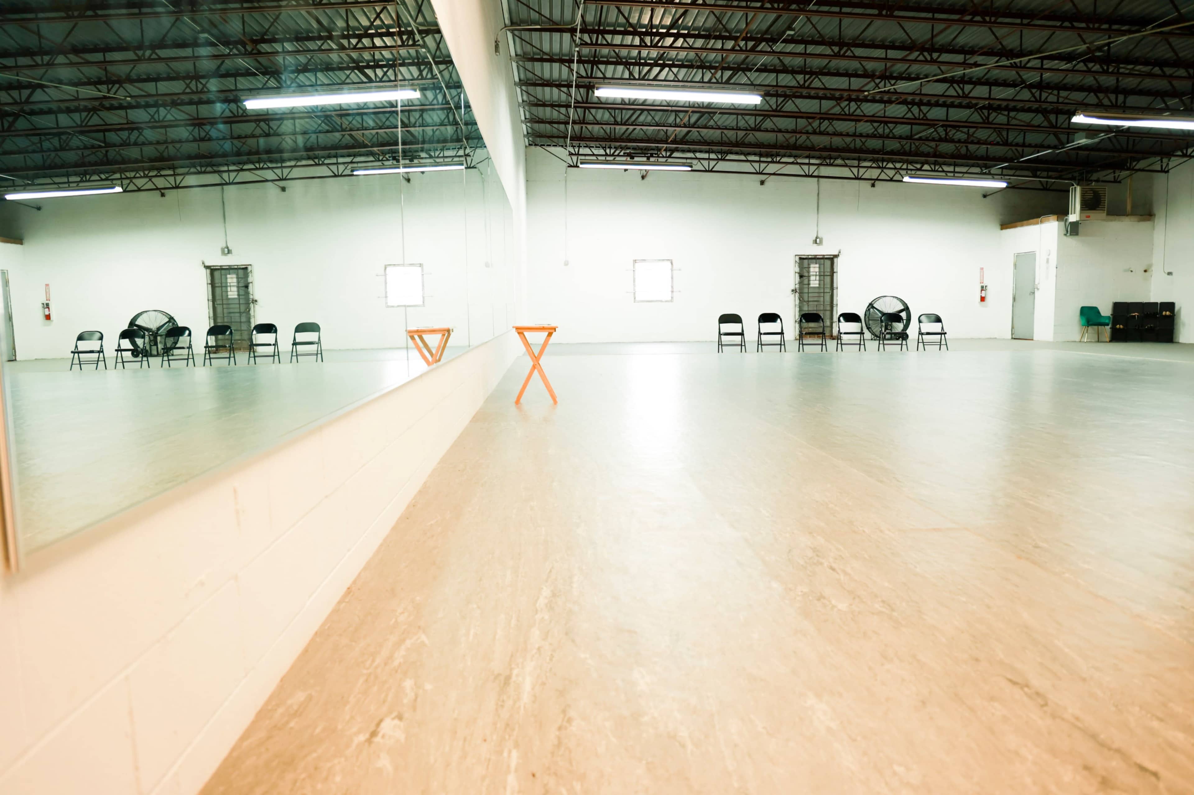 An empty room with wooden flooring, large mirrors on one wall, and a row of black chairs and fans along the opposite side.