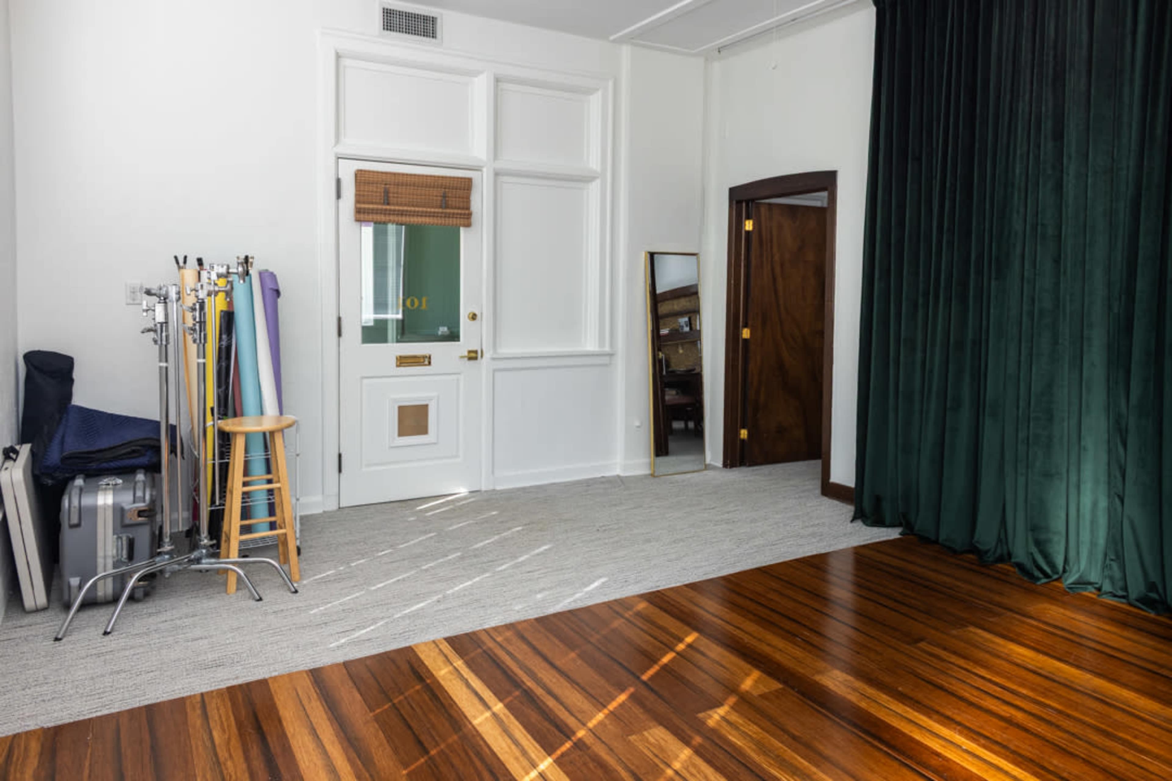 The image shows a spacious room with wooden flooring, a green curtain, a mirror, and photography equipment on a stand.