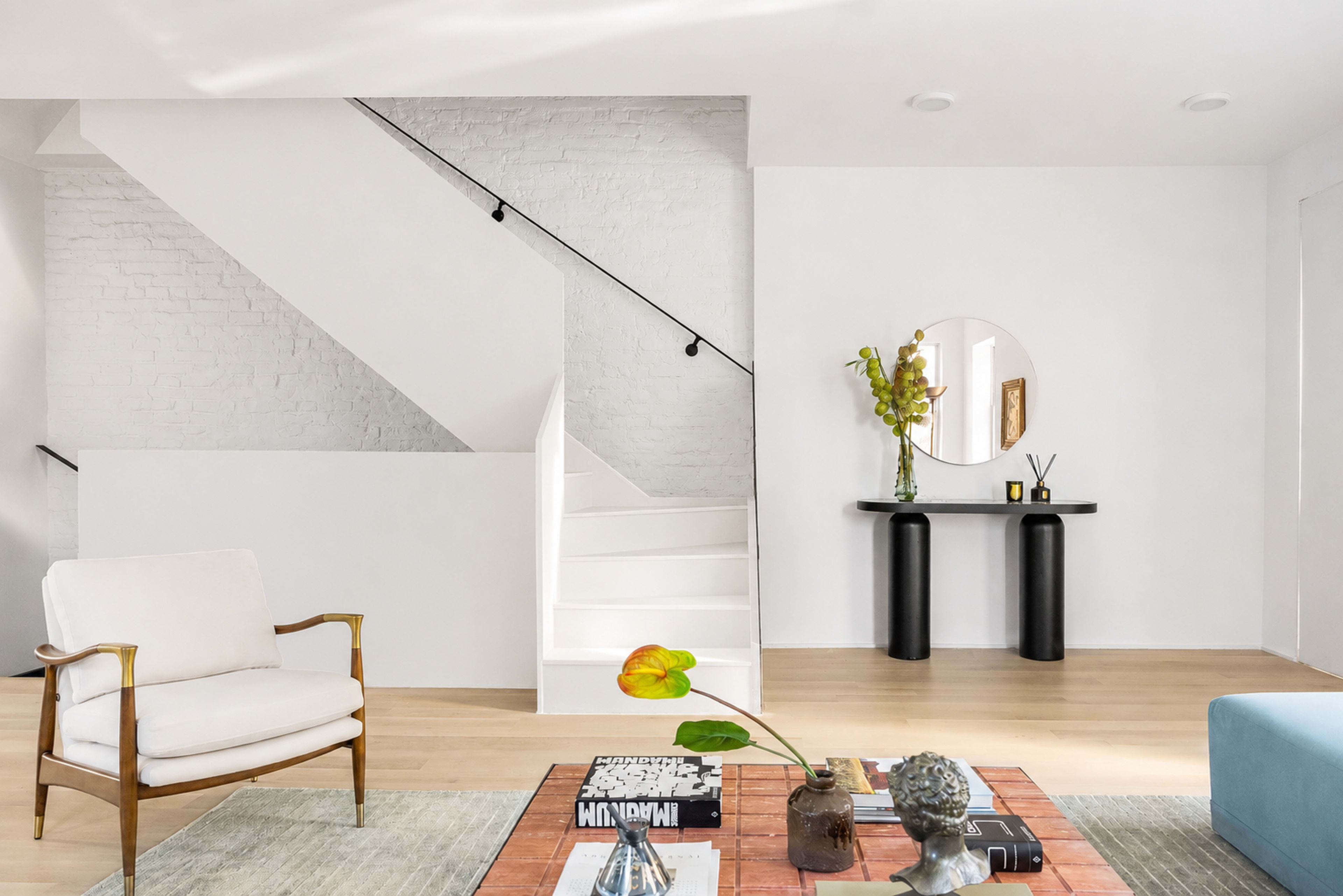 The image shows a modern interior living space with a white staircase, a circular wall mirror, a black console table, and a light-colored armchair.