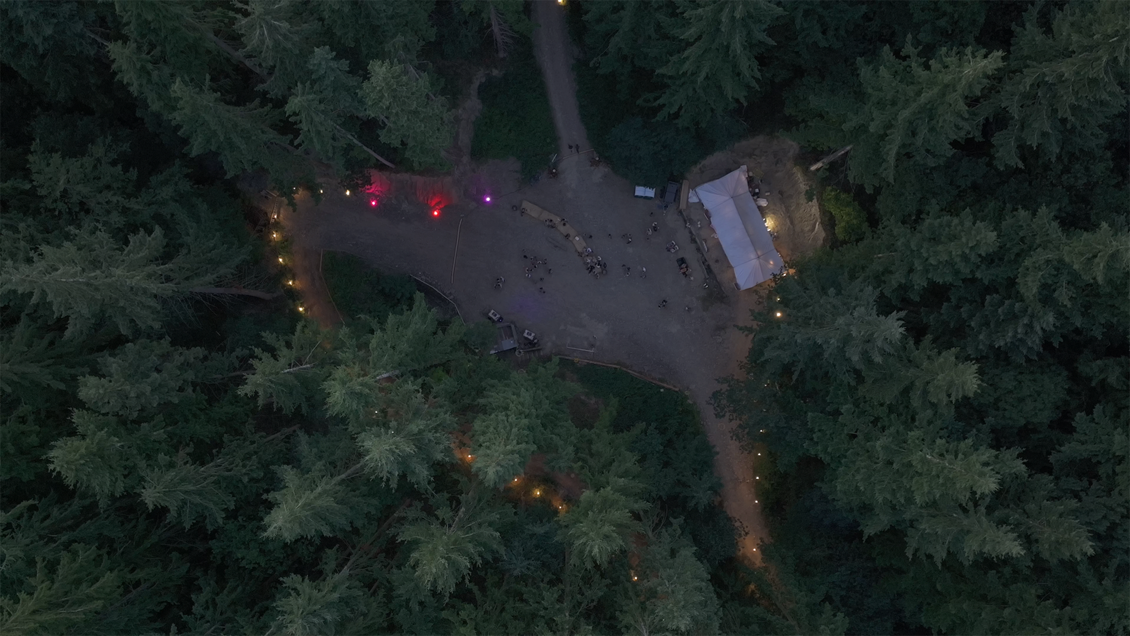 An aerial view shows a gathering area in a forested setting with a tent and pathways illuminated by lights among the tall trees.