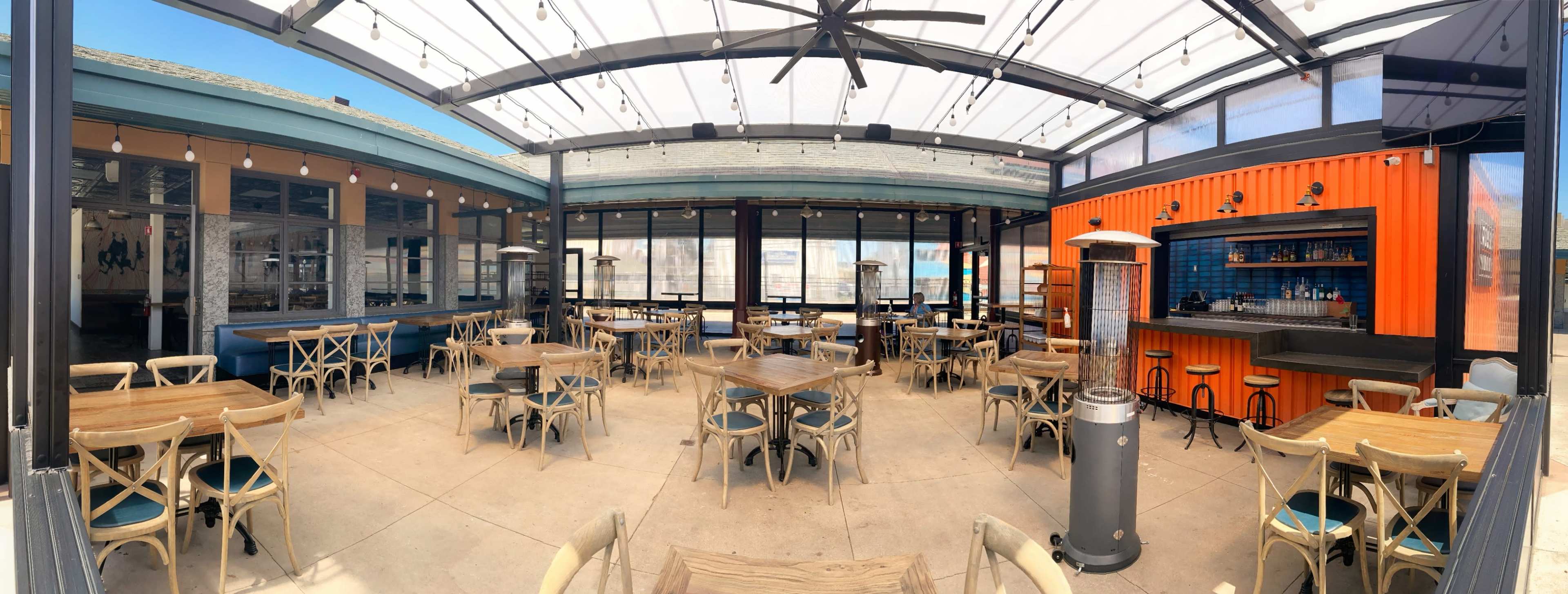 A spacious outdoor dining area with wooden tables and chairs, surrounded by large glass walls and a bar in an orange shipping container style.