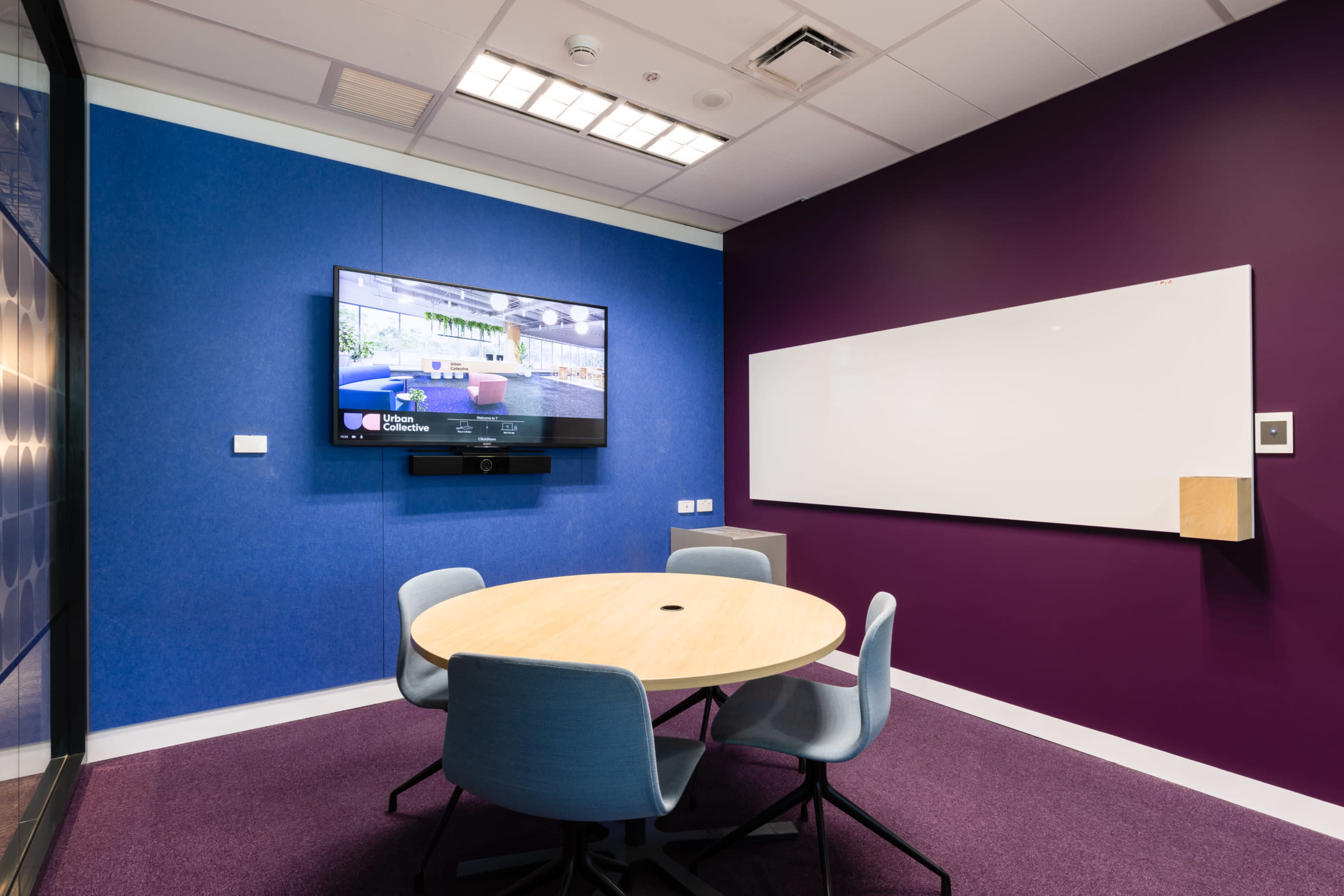 A small meeting room featuring a round table surrounded by four chairs, a wall-mounted television, and a whiteboard.