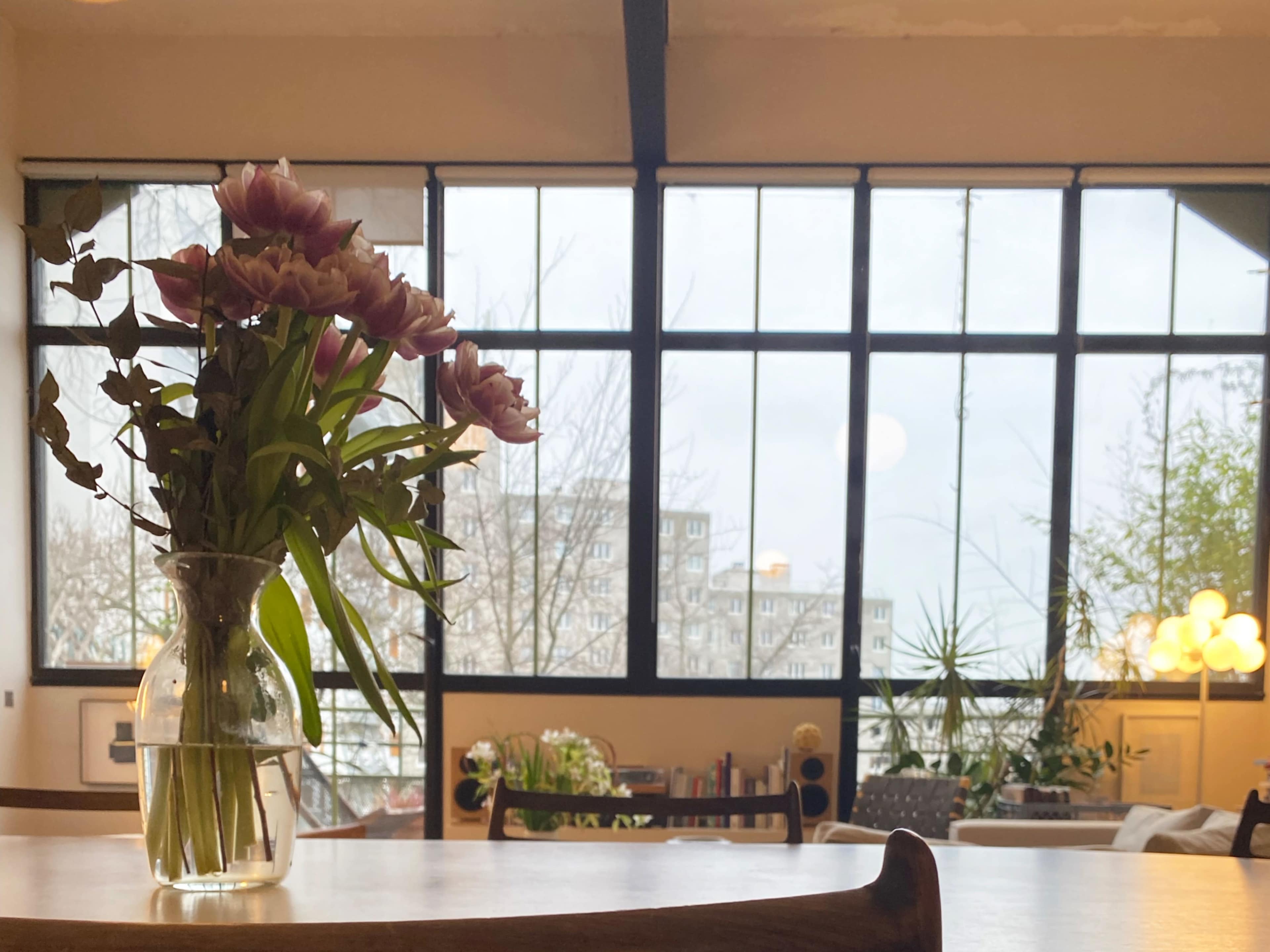 A vase of flowers is placed on a wooden table in a bright room with large windows that overlook a cityscape.