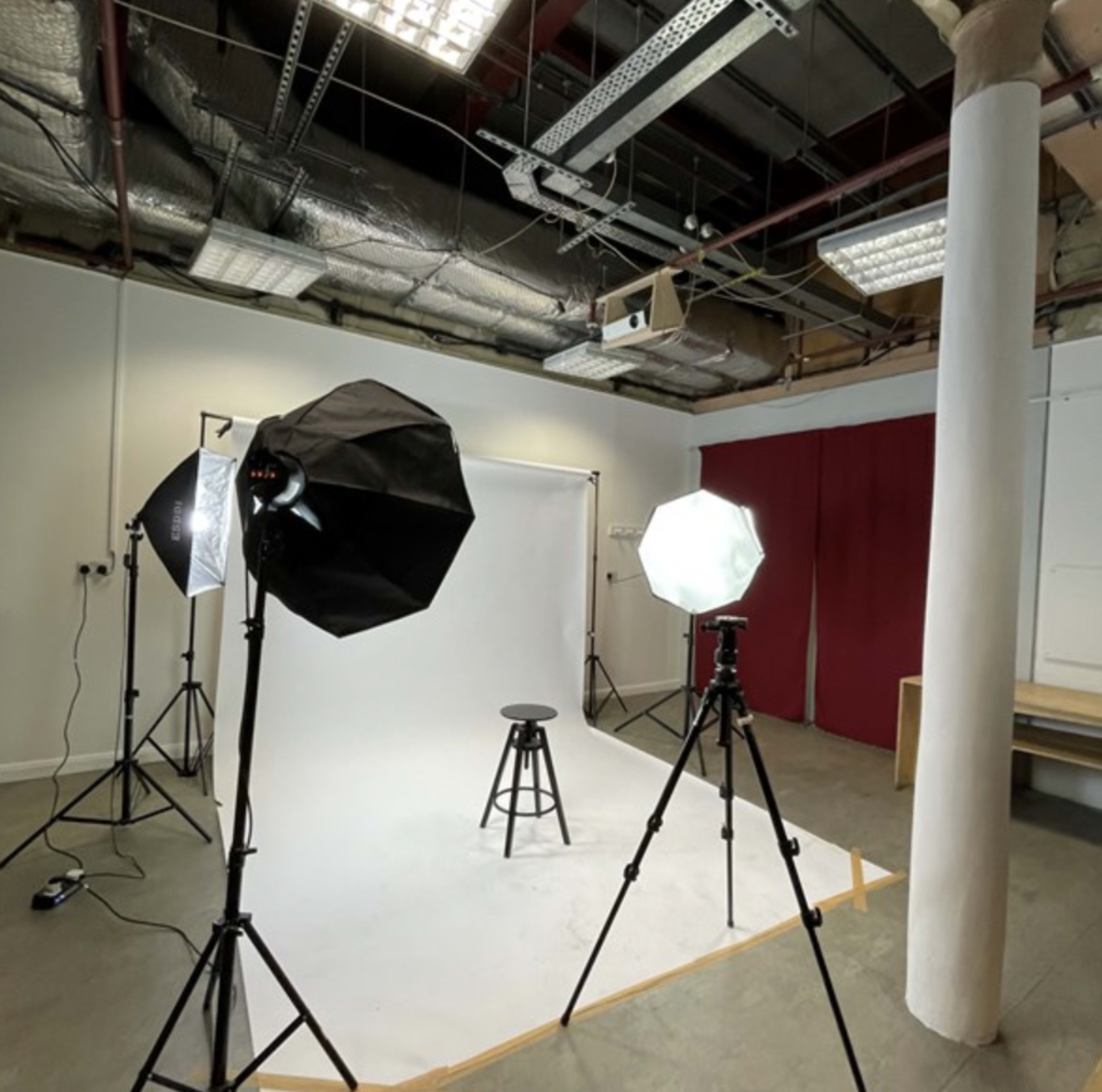 The image shows a photography studio equipped with softbox lights and a stool in front of a plain white backdrop.
