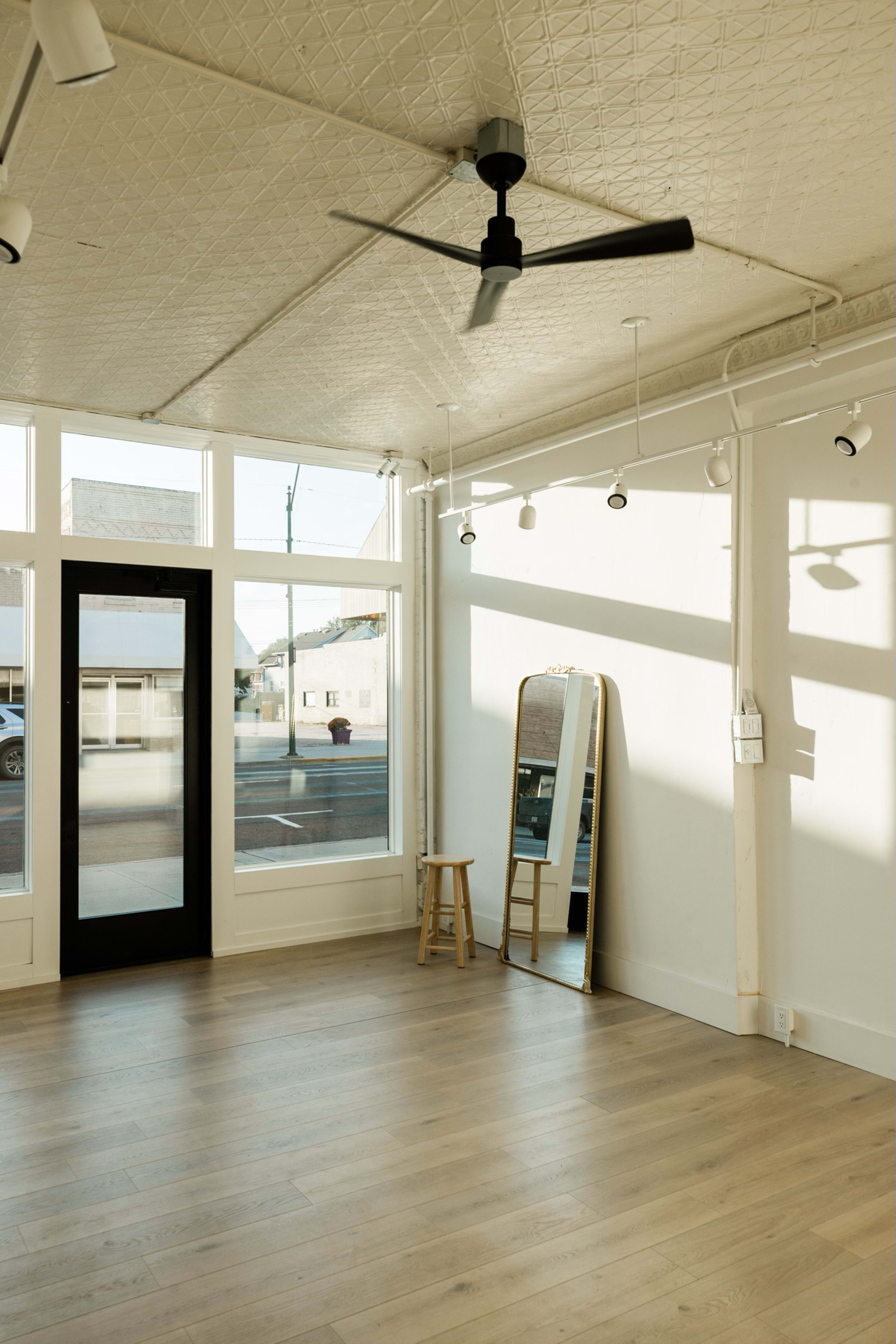 The image shows an empty room with large windows, a black door, a full-length mirror, a stool, and a ceiling fan.