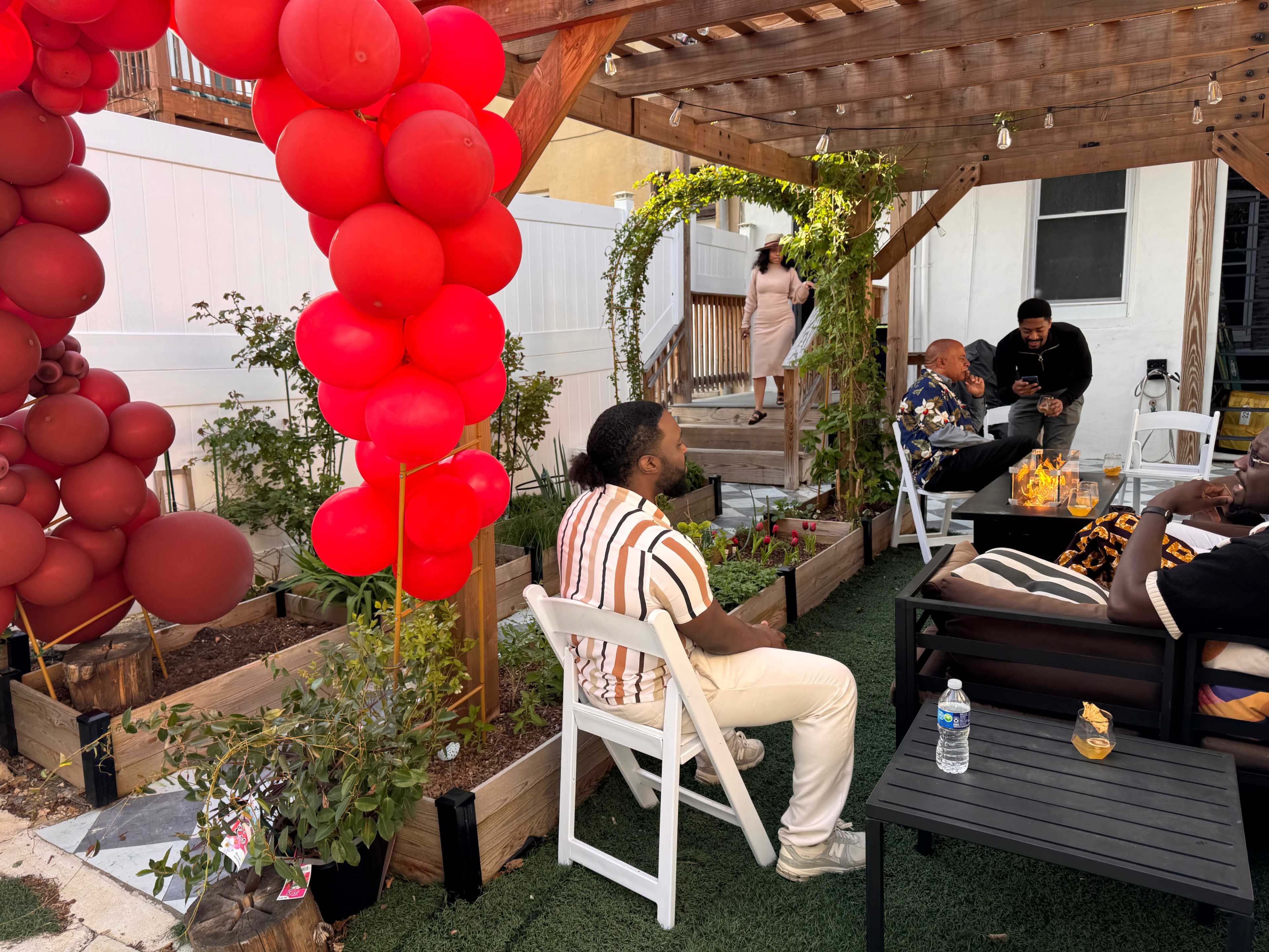 A man sits on a chair under a wooden pergola adorned with red balloons while others converse nearby in a garden setting.