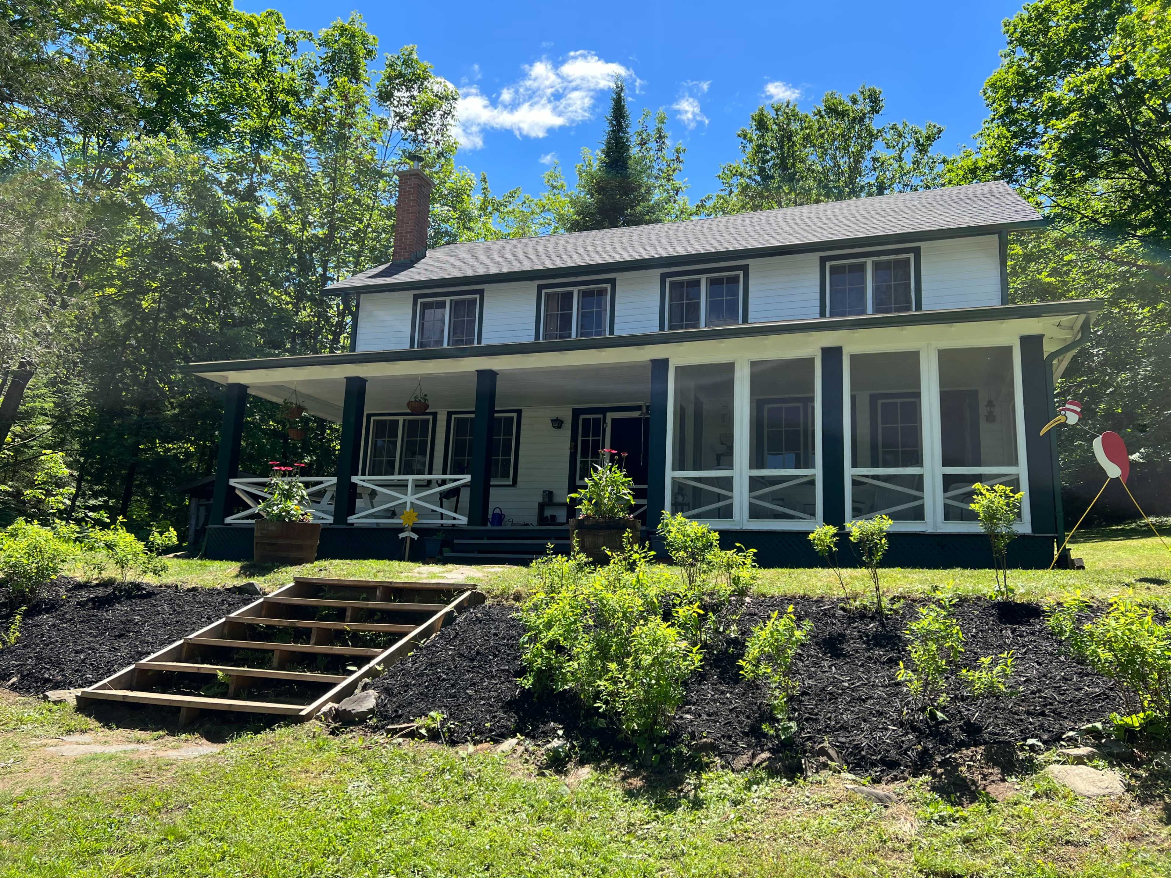 A two-story white house with dark green trim features a wide front porch, surrounded by trees and landscaped flower beds.