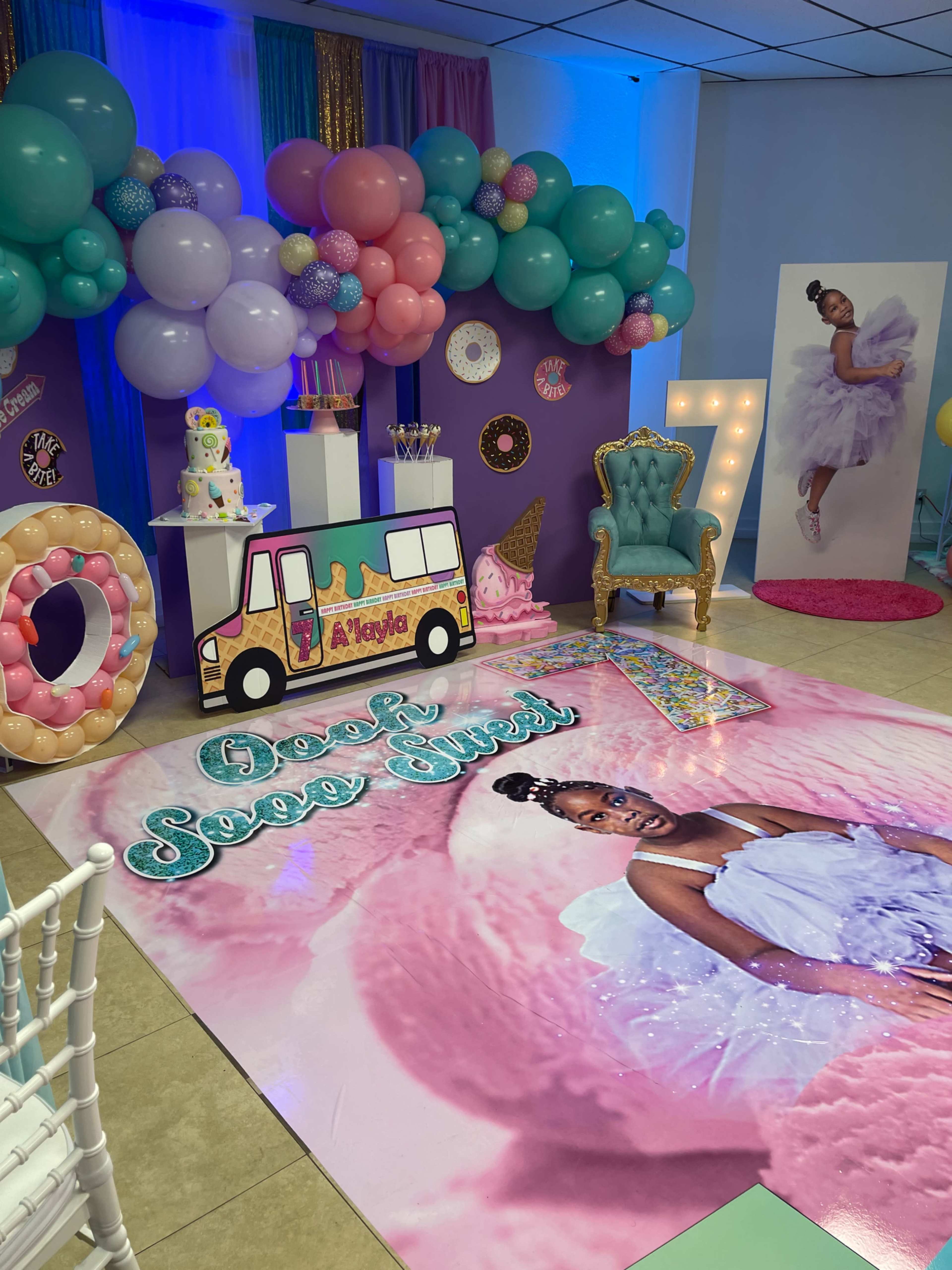 The scene features a colorful birthday celebration setup with balloons, a large printed mat of a girl in a dress, a dessert table, and decorative props.