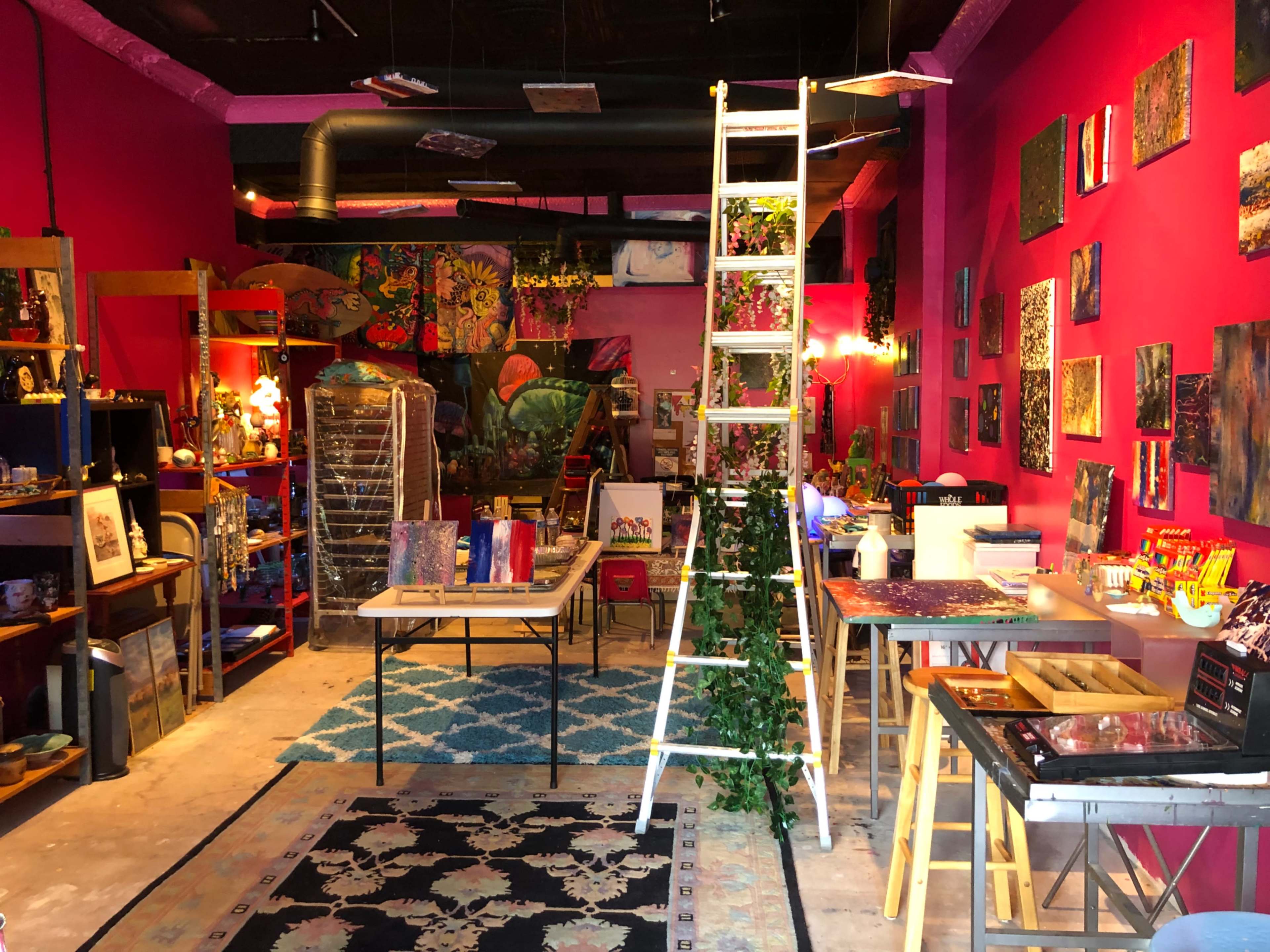 A colorful art studio with bright pink walls, various art supplies, and a ladder positioned in the center among tables and artworks.