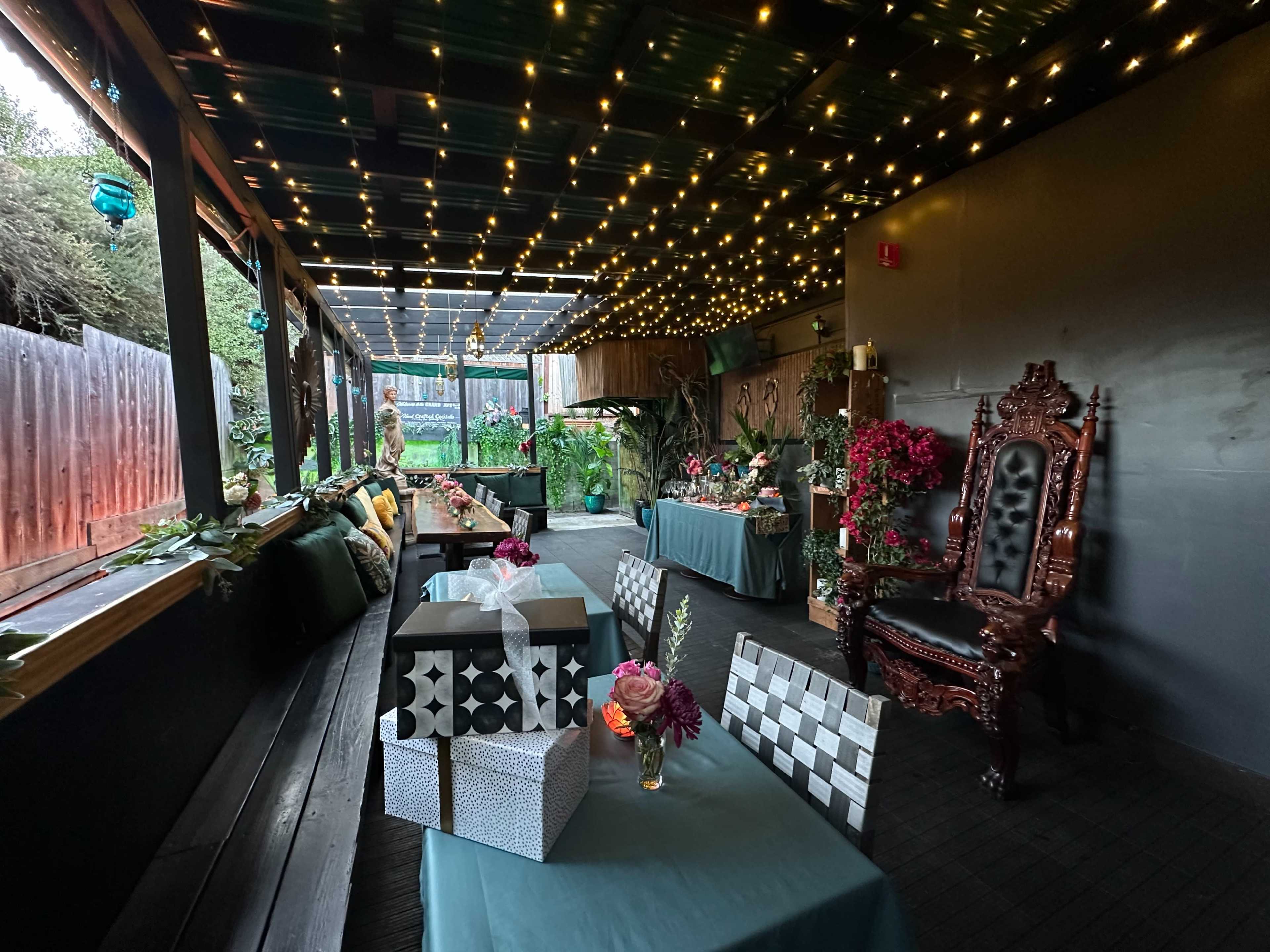 The image shows a decorated indoor space with festive lighting, dining tables covered in teal tablecloths, and various floral arrangements.