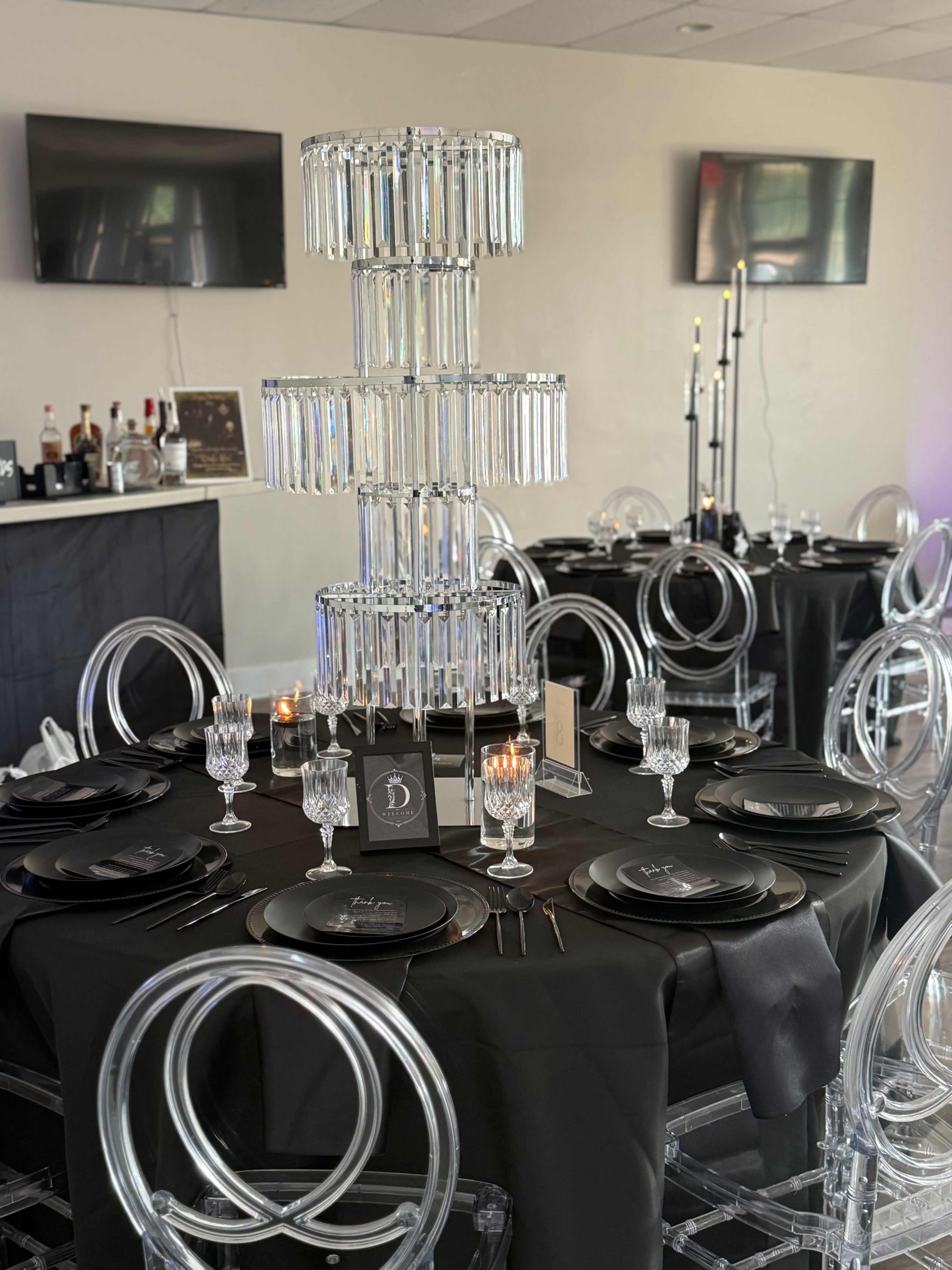 A dining table is elegantly set with black plates and crystal glassware, featuring a tall, decorative centerpiece made of glass and arranged in a modern venue.