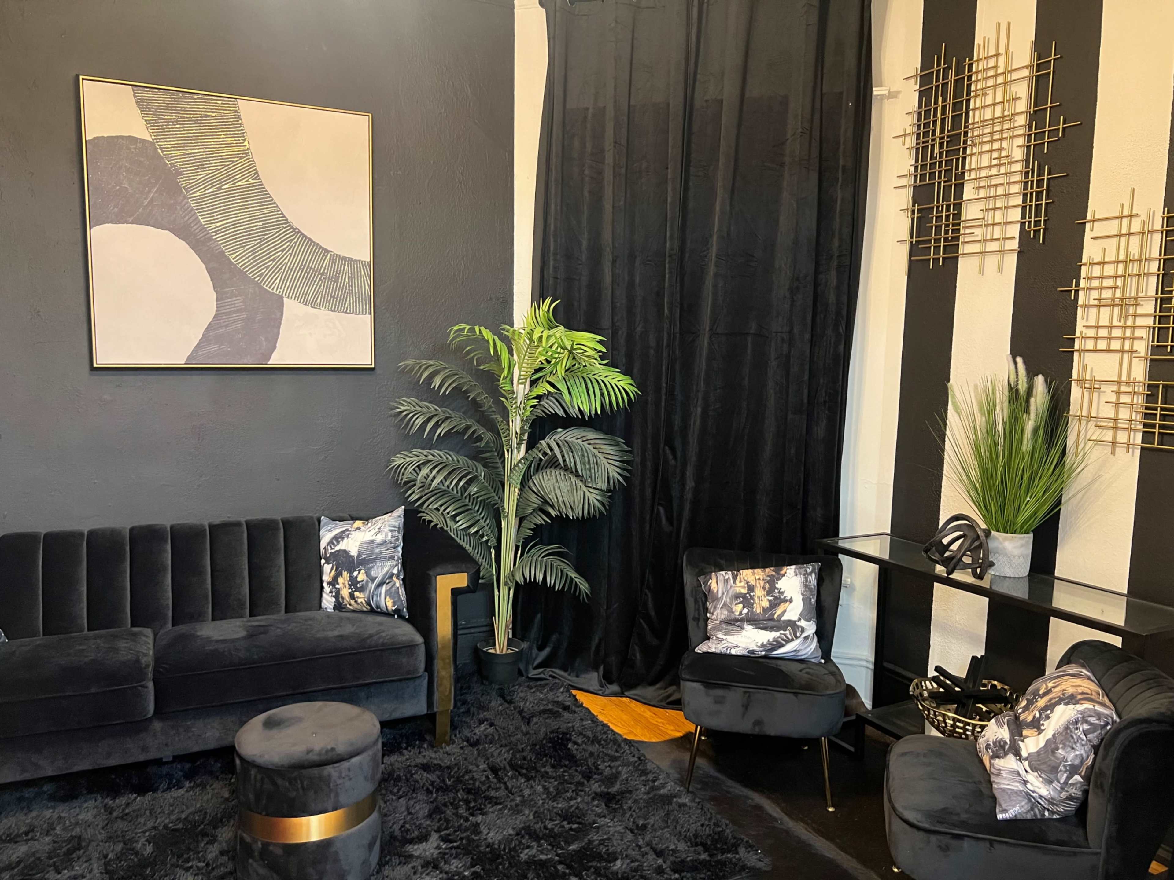 The image shows a modern living room with a black velvet sofa, a round ottoman, a glass table, and a tall green plant against a backdrop of black and white striped walls.