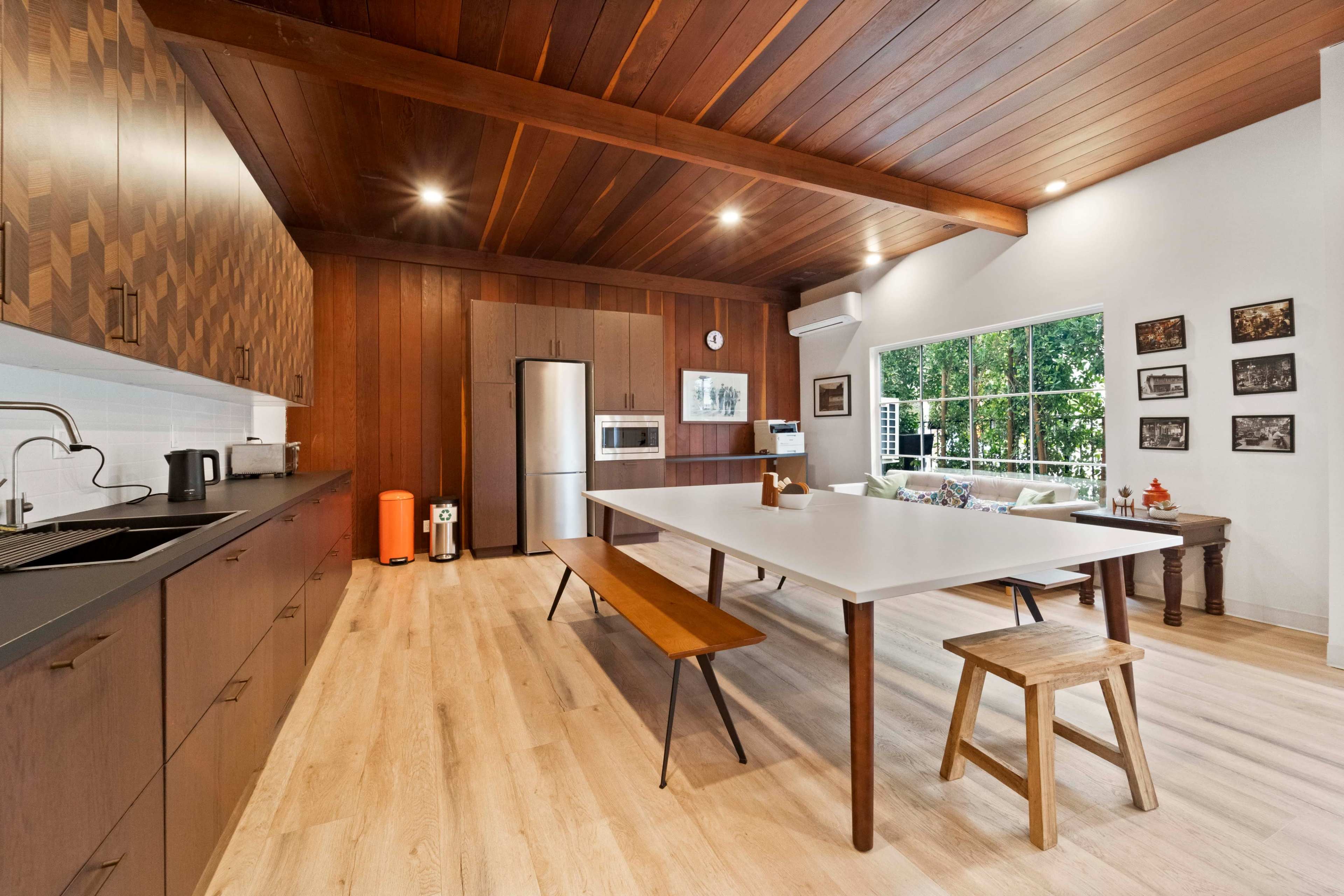 A modern kitchen features a large white table, wooden cabinetry, and a view of greenery through a large window.