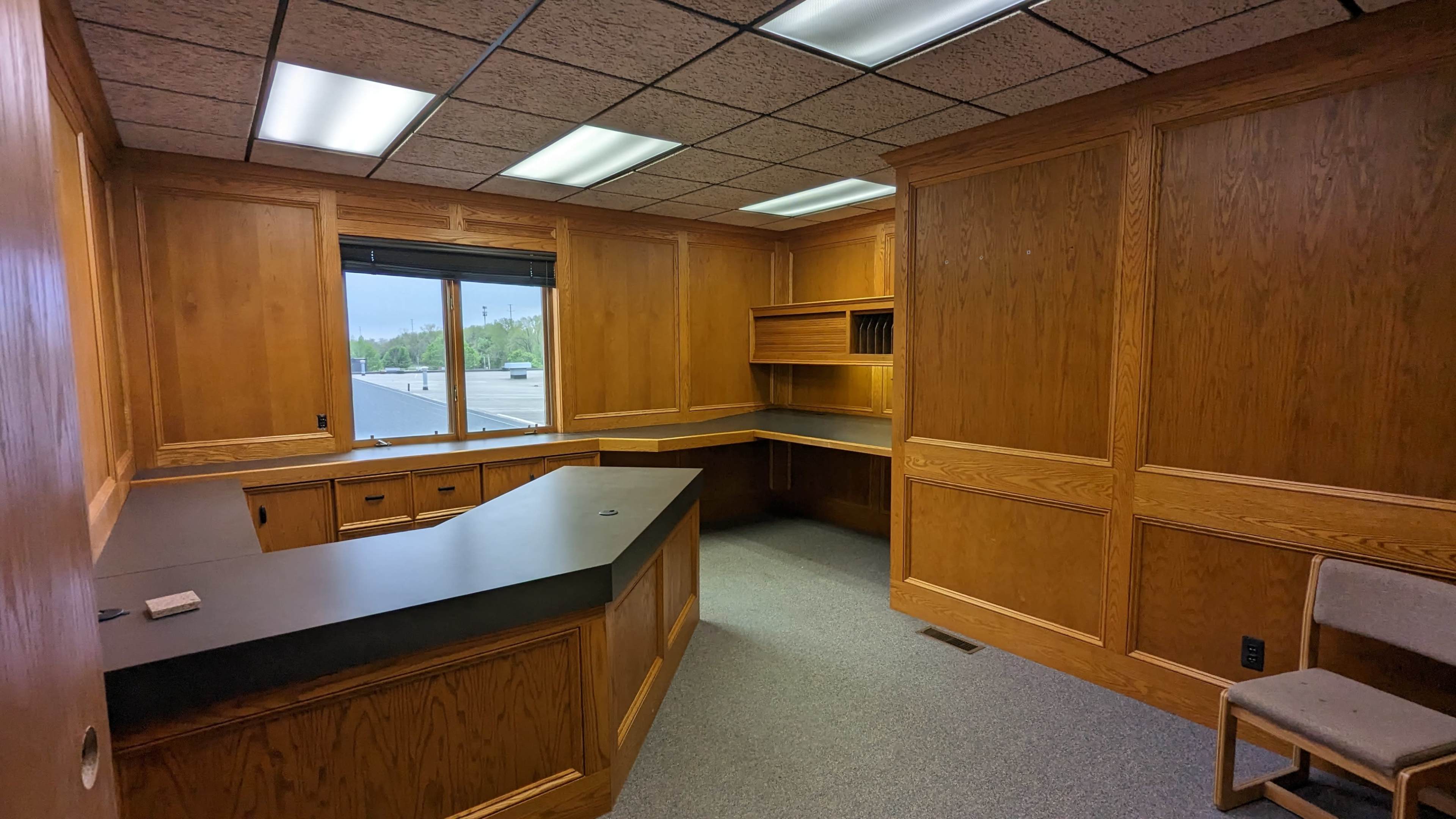 A vacant office space with wooden paneling, a large desk, and a window overlooking an external view.