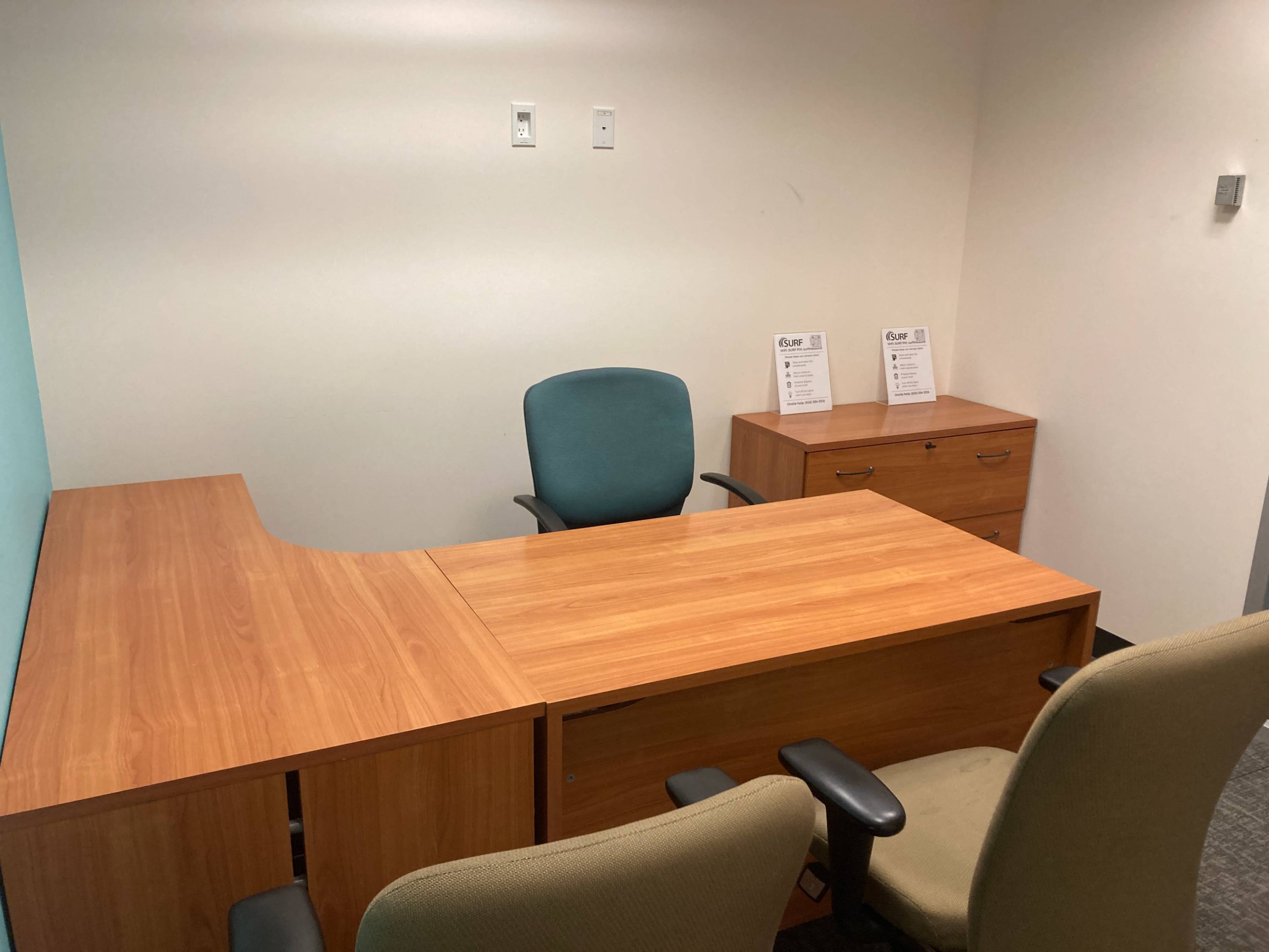 A minimalist office setup with a wooden desk, two chairs, and a filing cabinet against a wall.