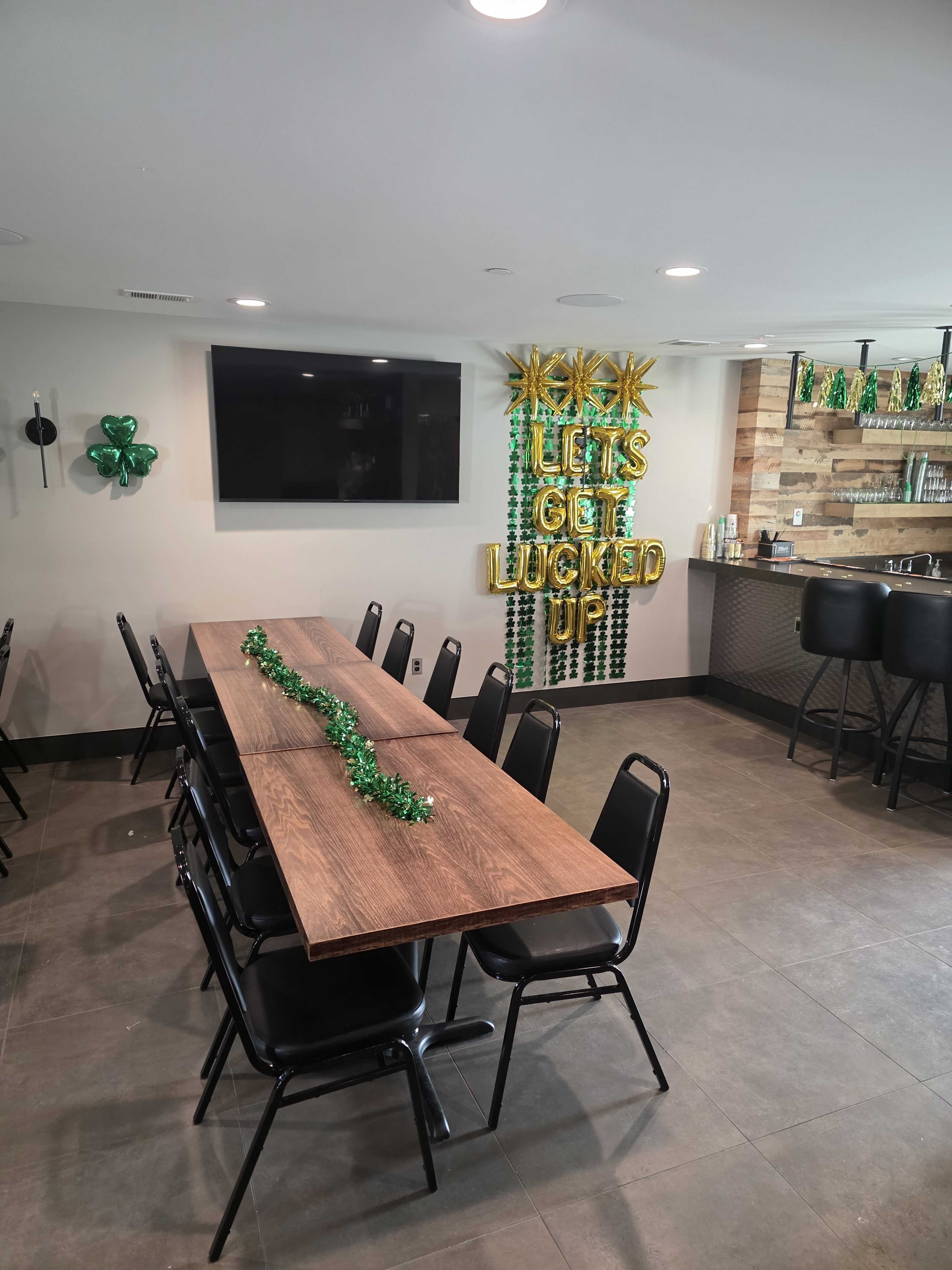 The image shows a modern gathering space with a long wooden table surrounded by black chairs, decorated for a celebration with green decorations and a sign that reads "LET'S GET LUCKED UP."
