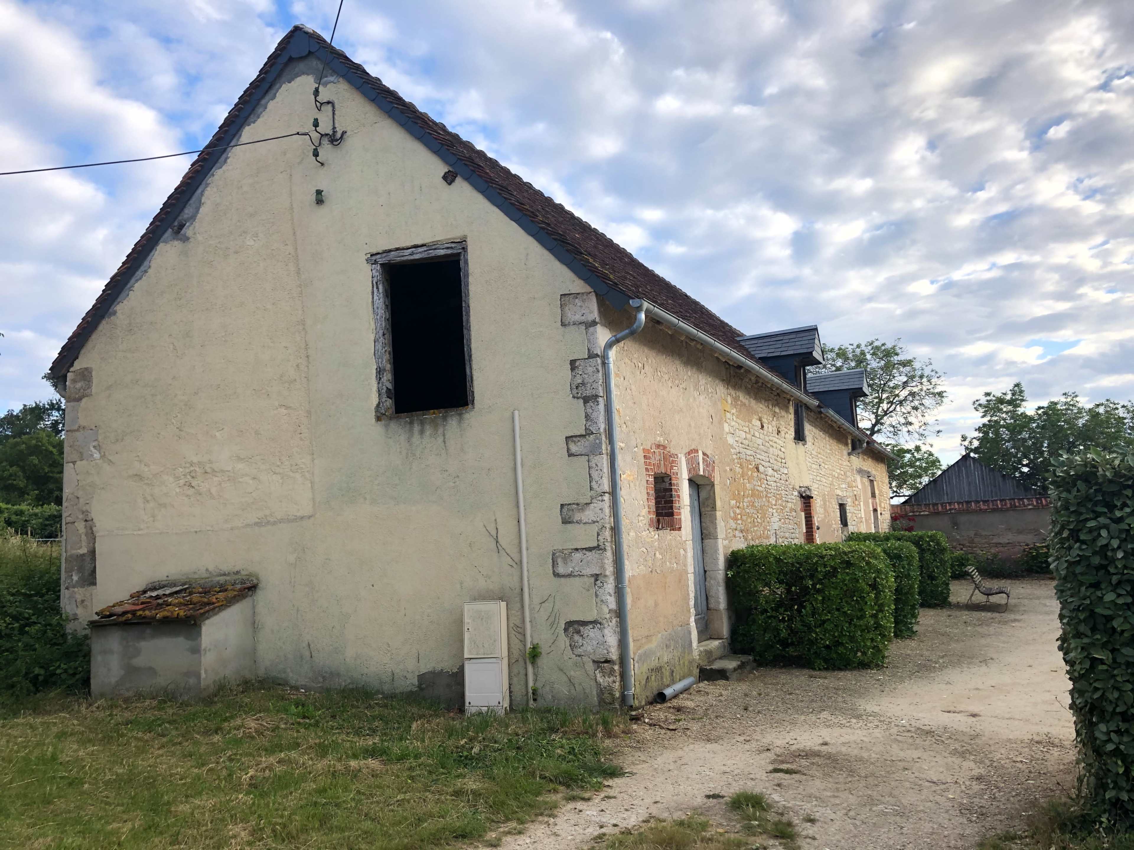The image shows an old, weathered house with a partially open window, surrounded by greenery and a gravel pathway.