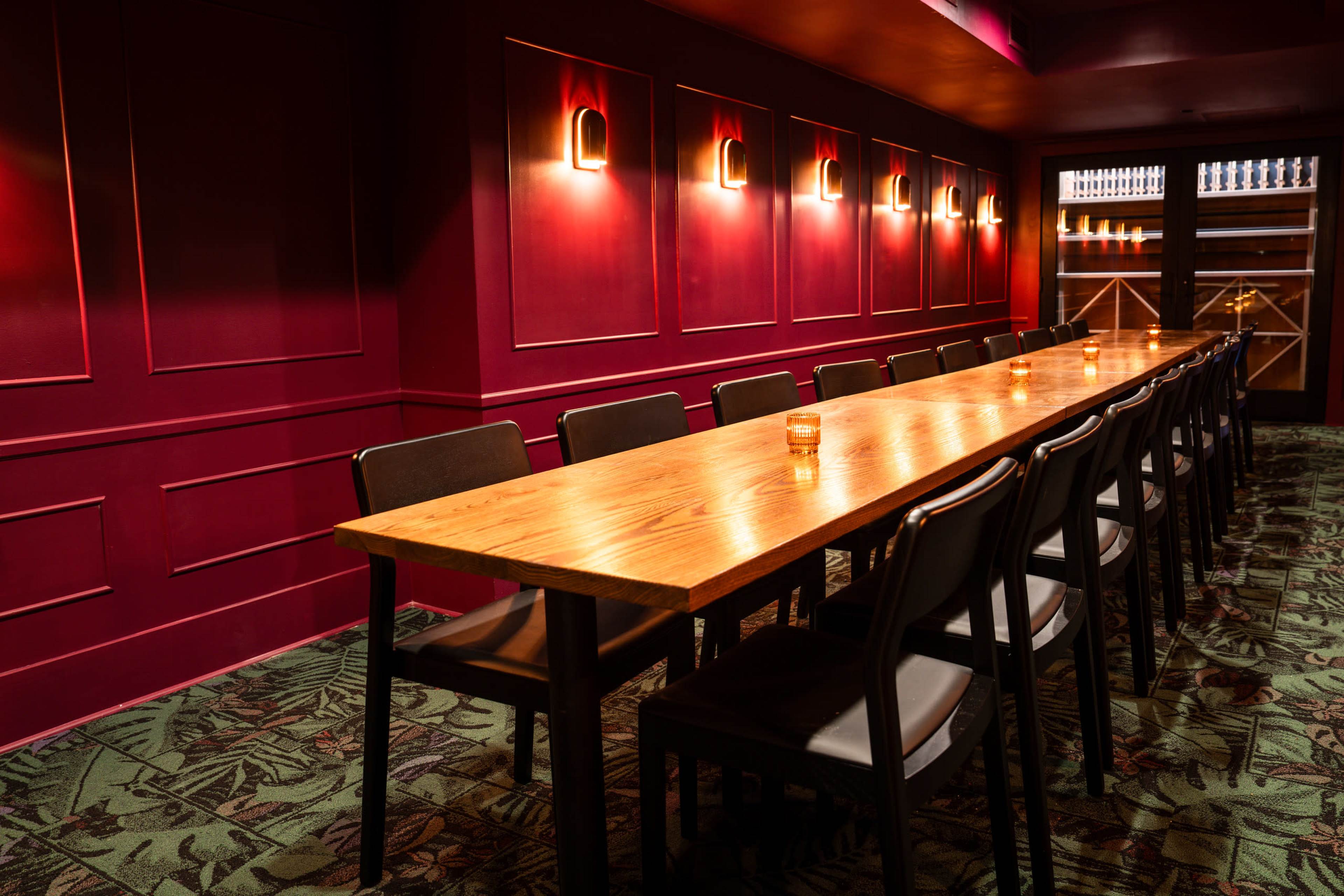 A long wooden table is set in a dimly lit room with purple walls, surrounded by chairs and illuminated by wall sconces.