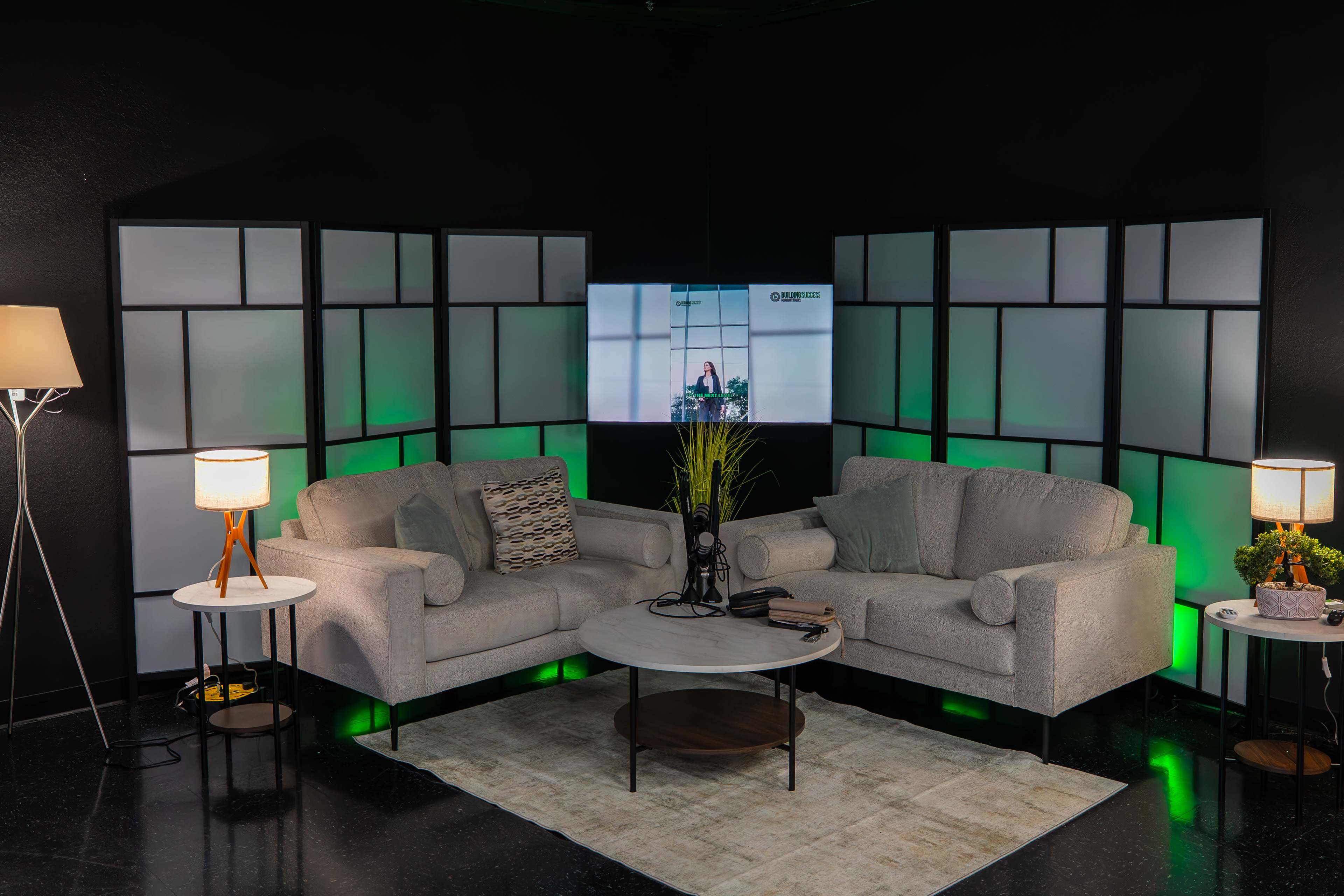 The image shows a modern studio setup featuring two light gray sofas, a round coffee table, and decorative lamps, set against a black wall with green-lit screens.