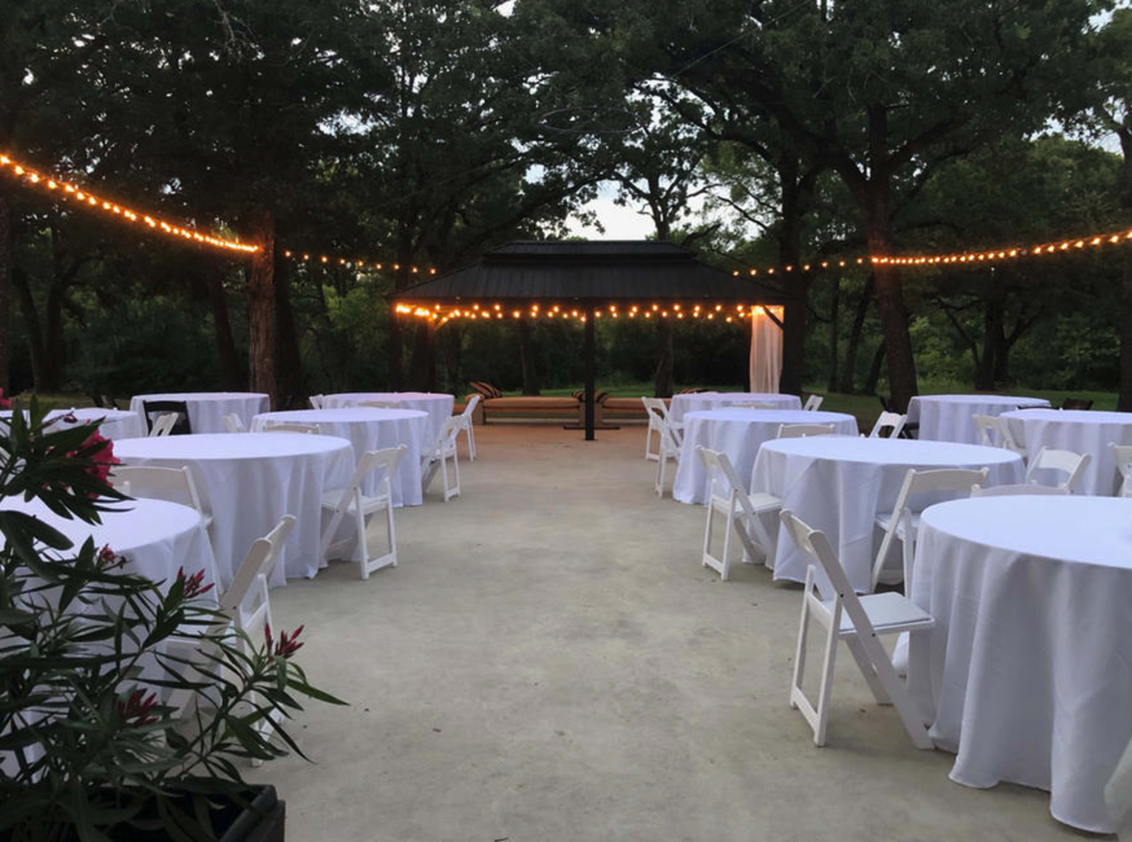 A landscaped outdoor area is set up for an event with round tables covered in white linens and string lights overhead.