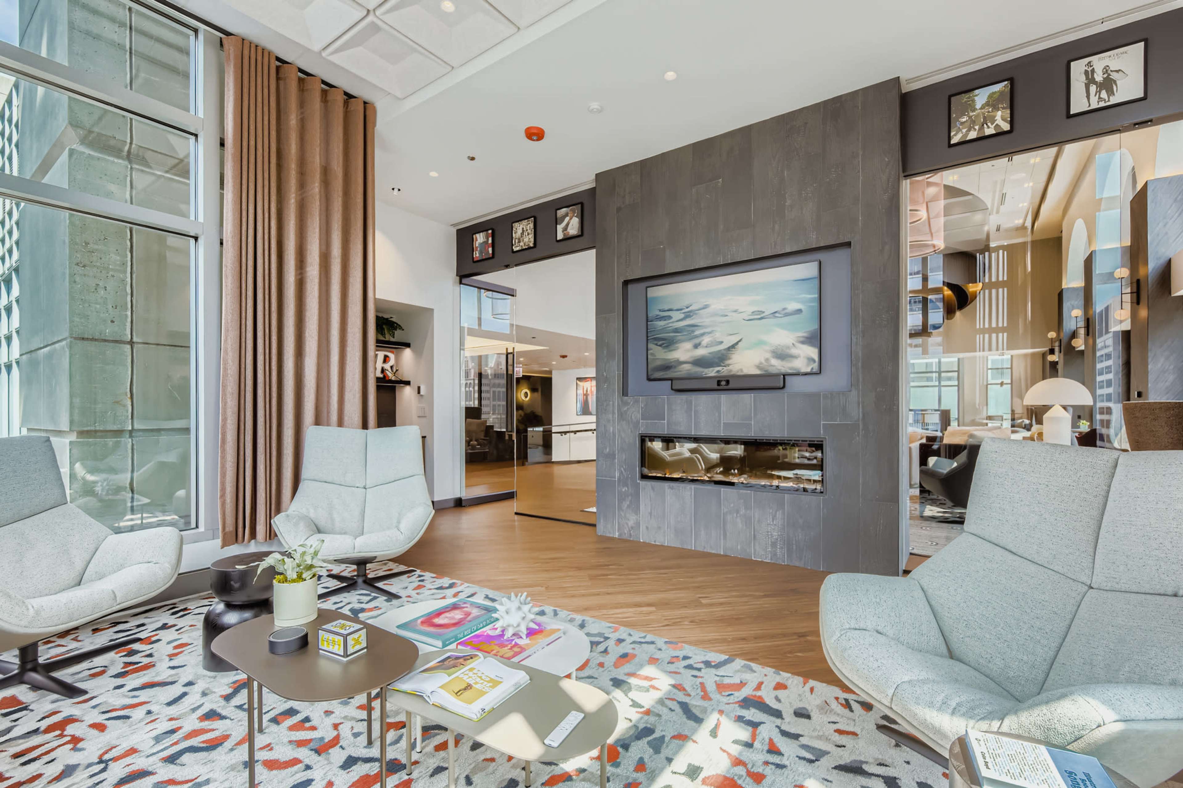 The image shows a modern living space featuring two light blue armchairs facing a low table, with a wall-mounted television above a sleek fireplace and large windows providing natural light.