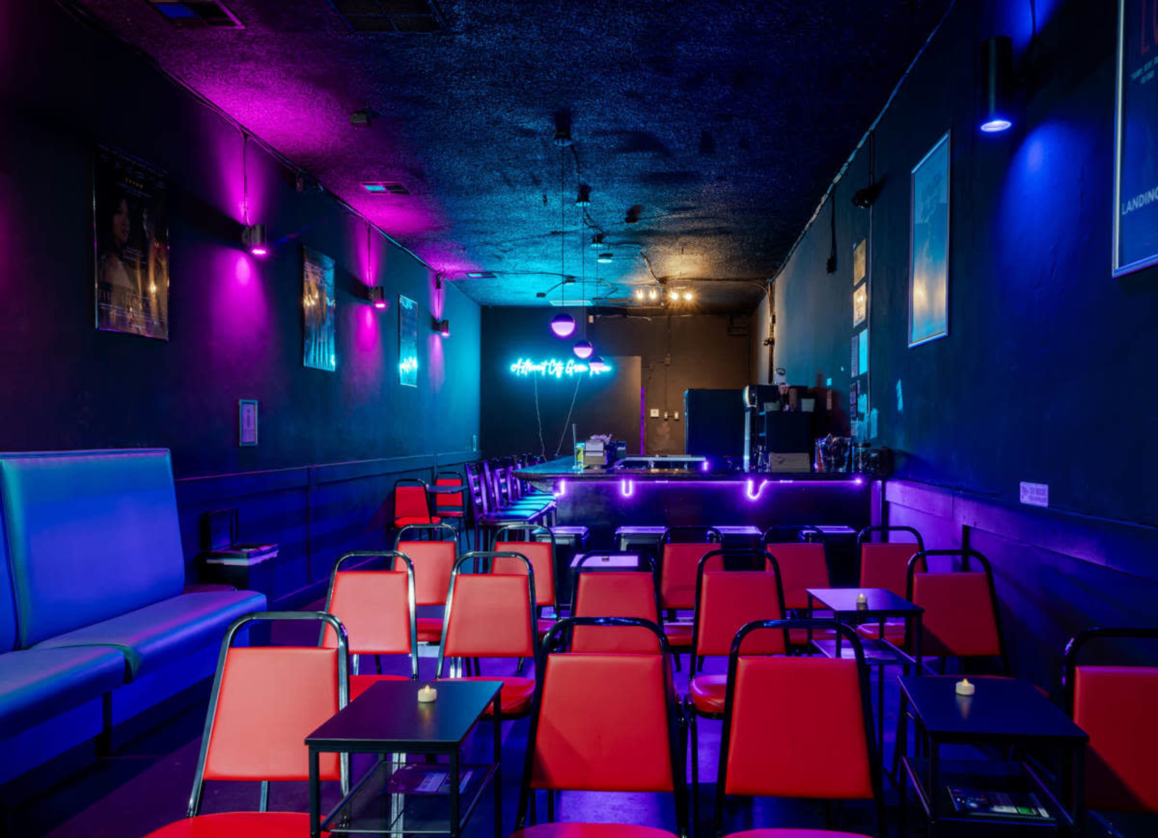 The image shows an empty room with rows of red chairs and small tables, illuminated by purple and blue lighting, featuring a bar at the back.