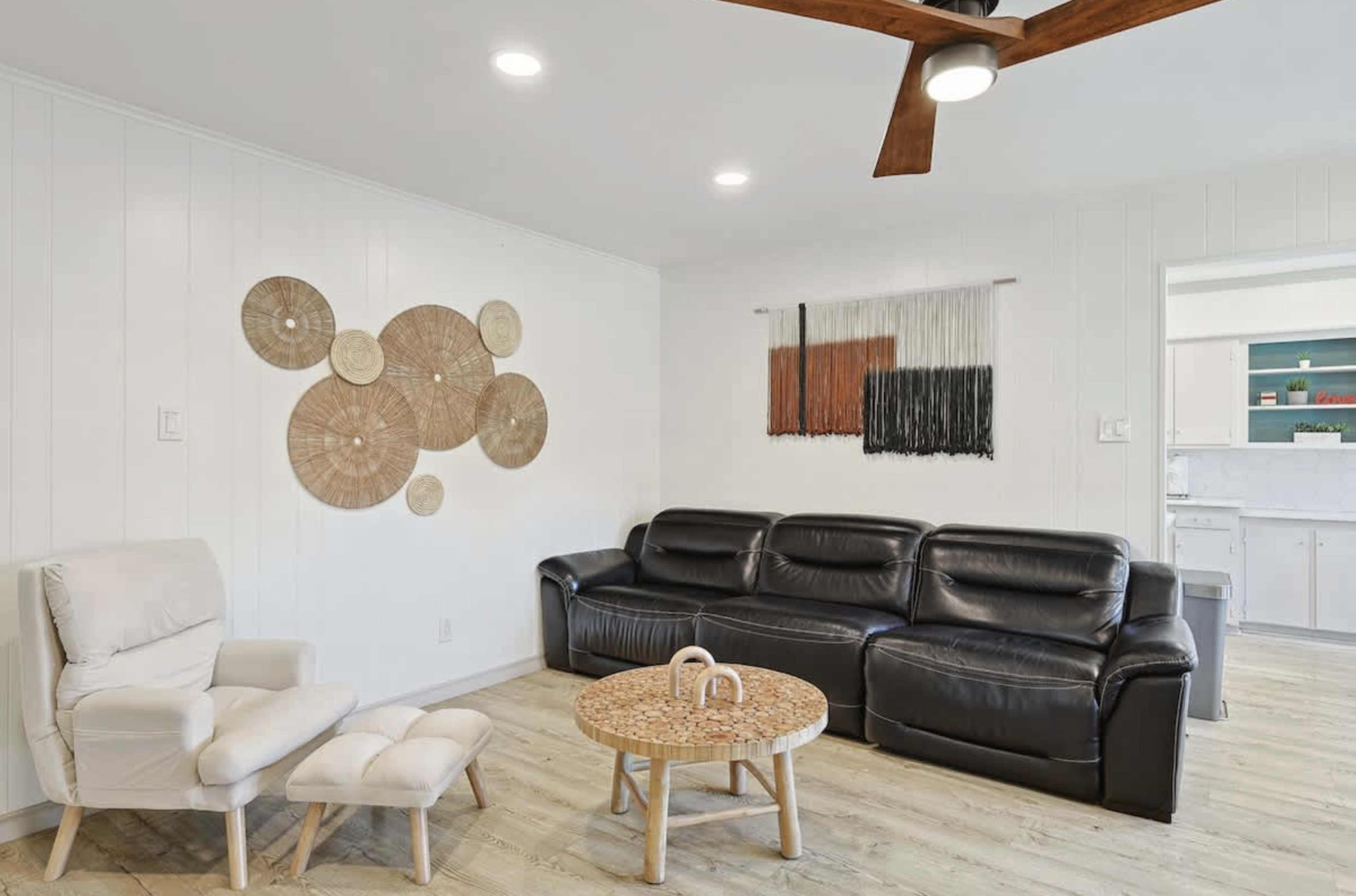 The image shows a modern living room with a black leather sofa, a light wooden floor, a round coffee table, and decorative wall hangings.