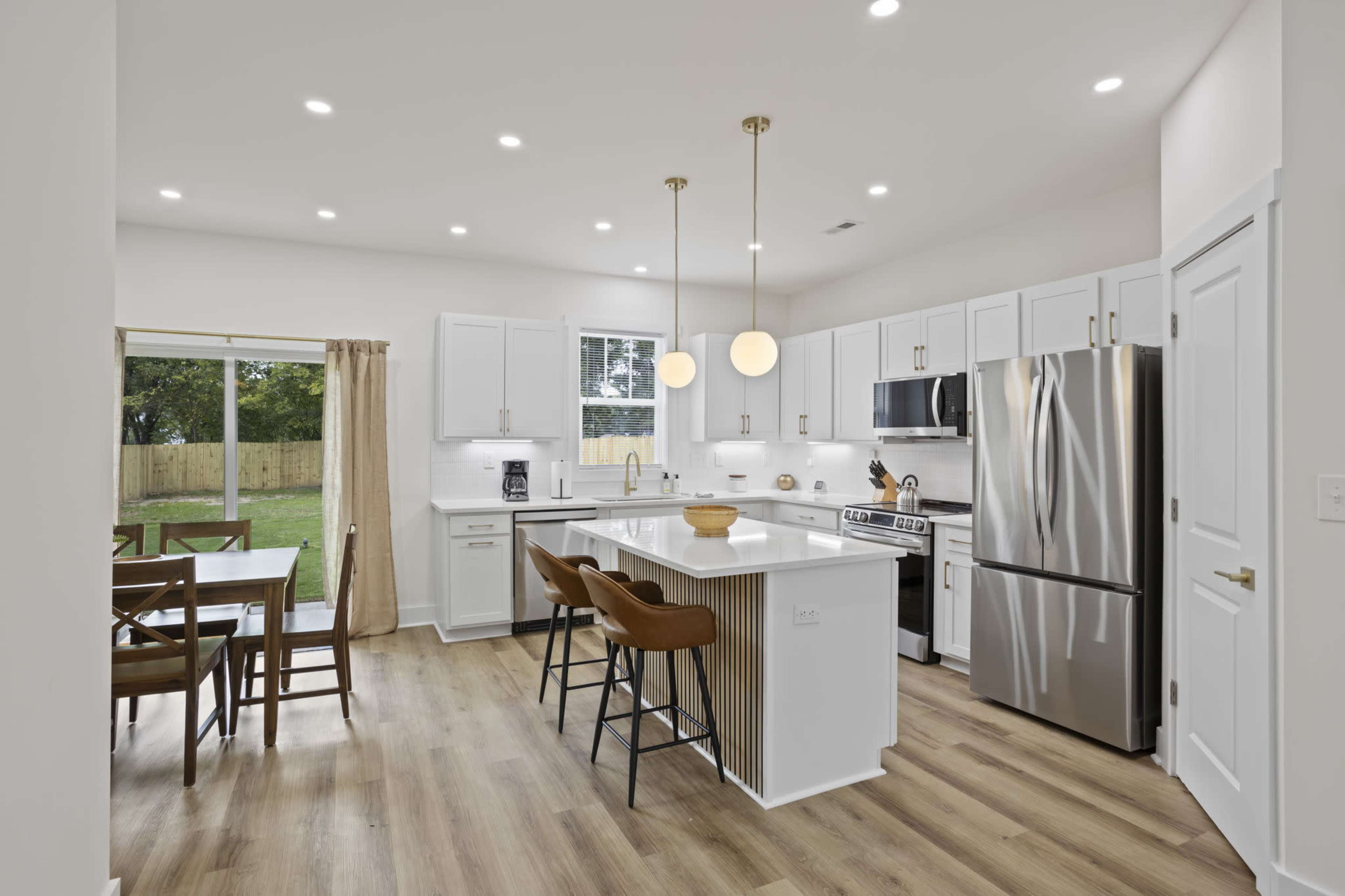 A modern kitchen features white cabinets, stainless steel appliances, and an island with pendant lights, alongside a dining area and large windows overlooking a backyard.