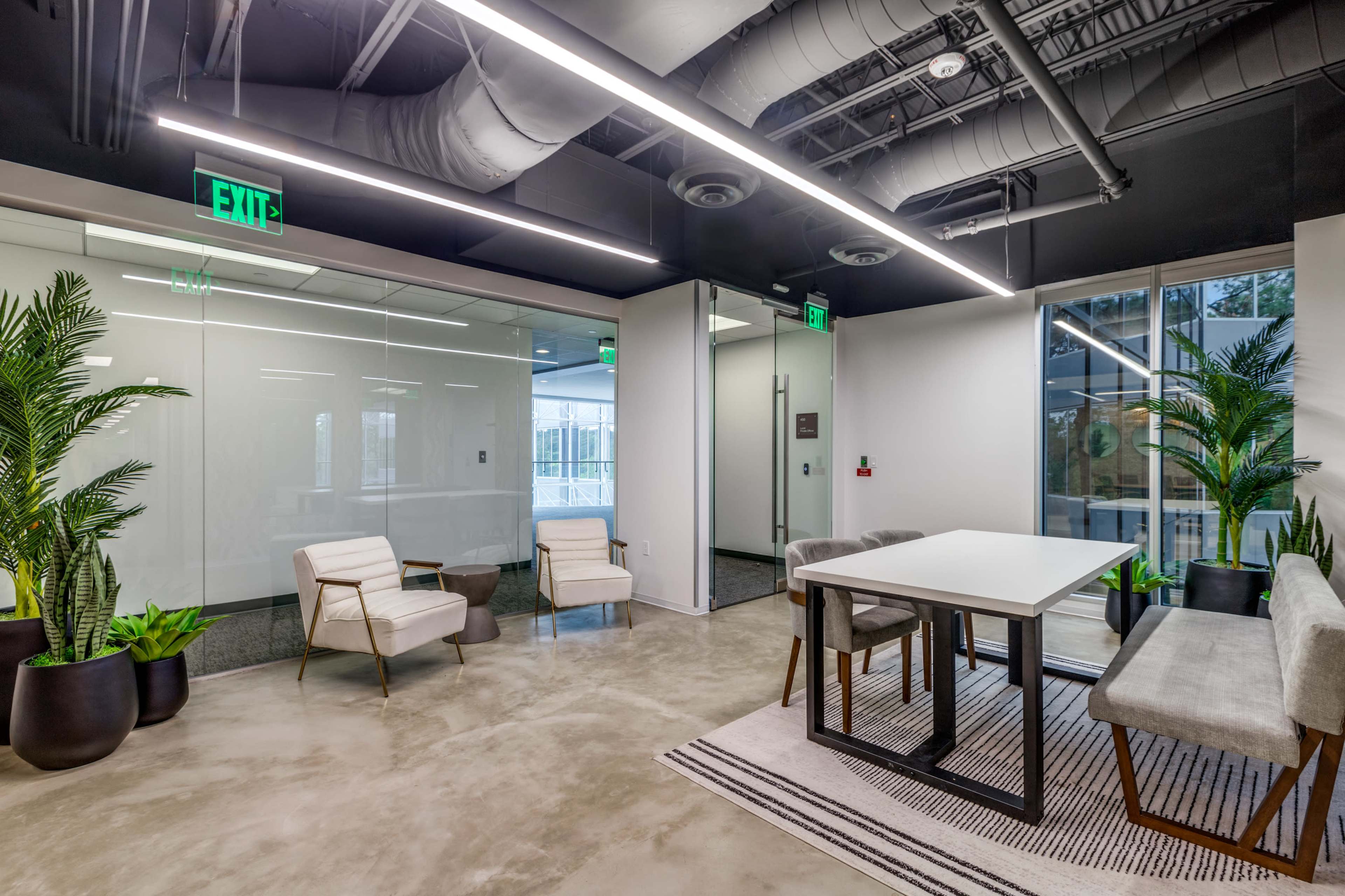 The image shows a modern office space featuring a seating area with two chairs, a dining table surrounded by gray chairs, large windows, and indoor plants.