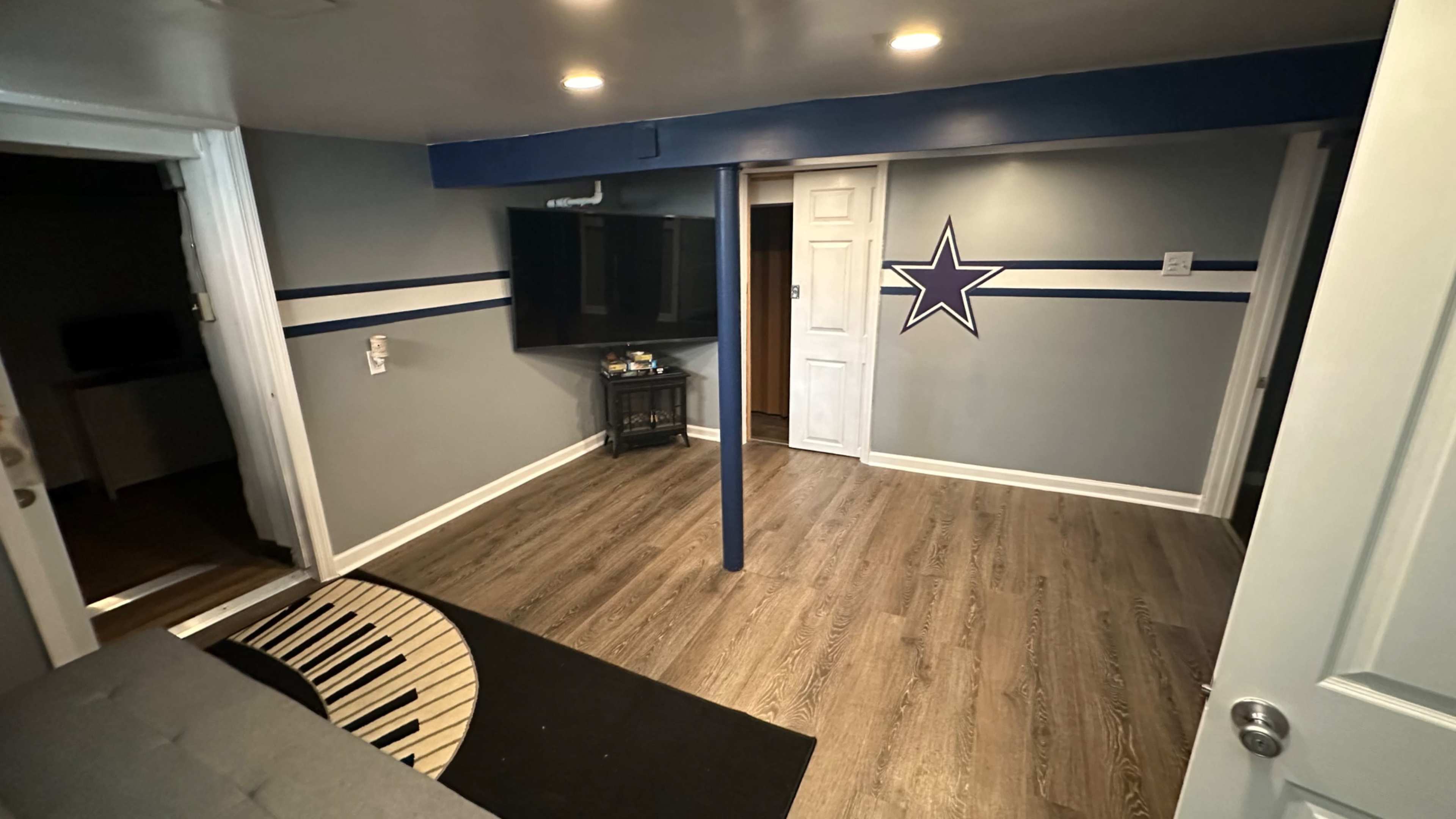 A basement room with brown wooden flooring, a television mounted on the wall, and a star decoration, featuring blue and white stripes.