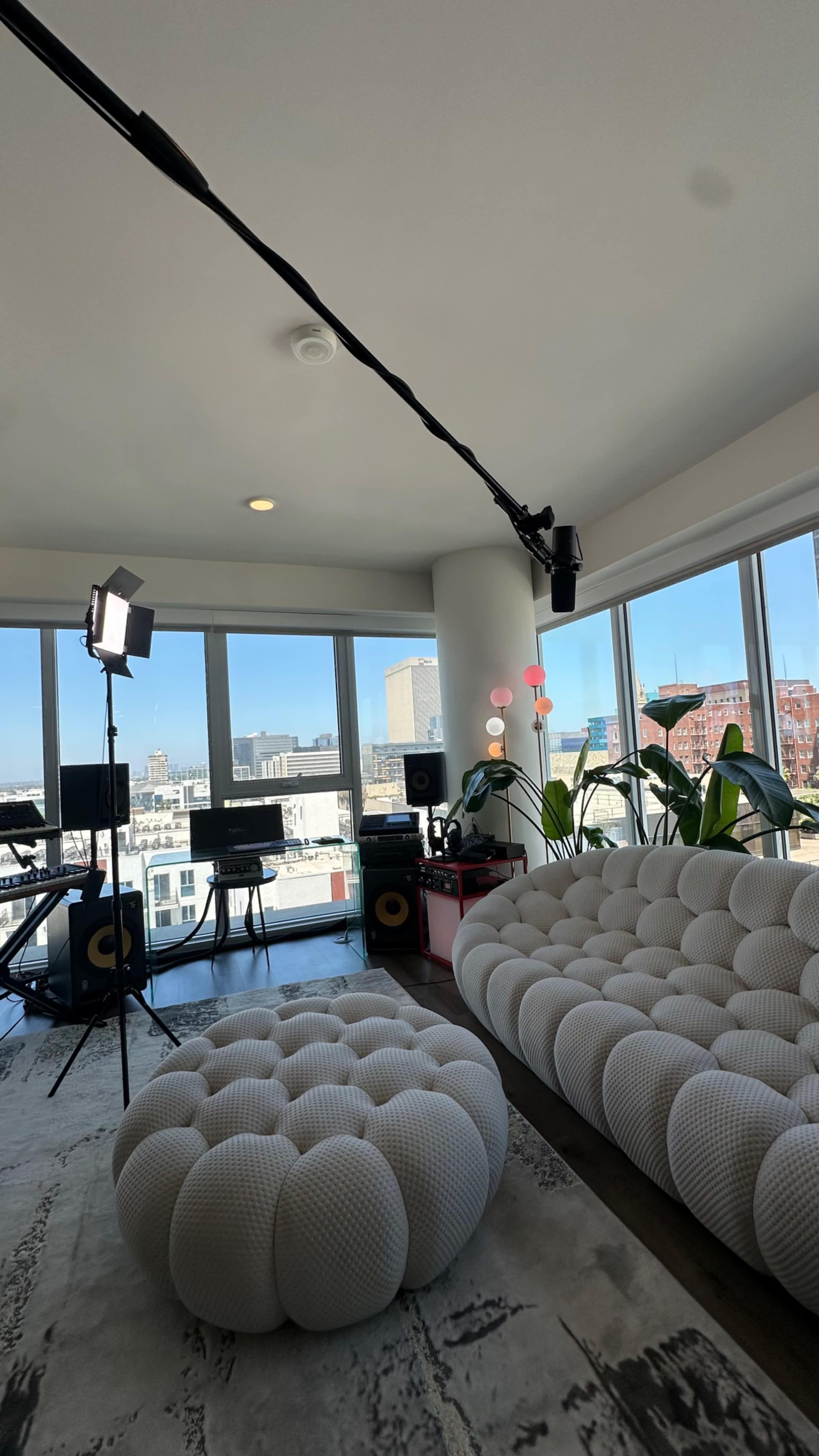 The image shows a modern, bright living room with large windows, a minimalist seating area featuring round sofas, and a music setup in the background.