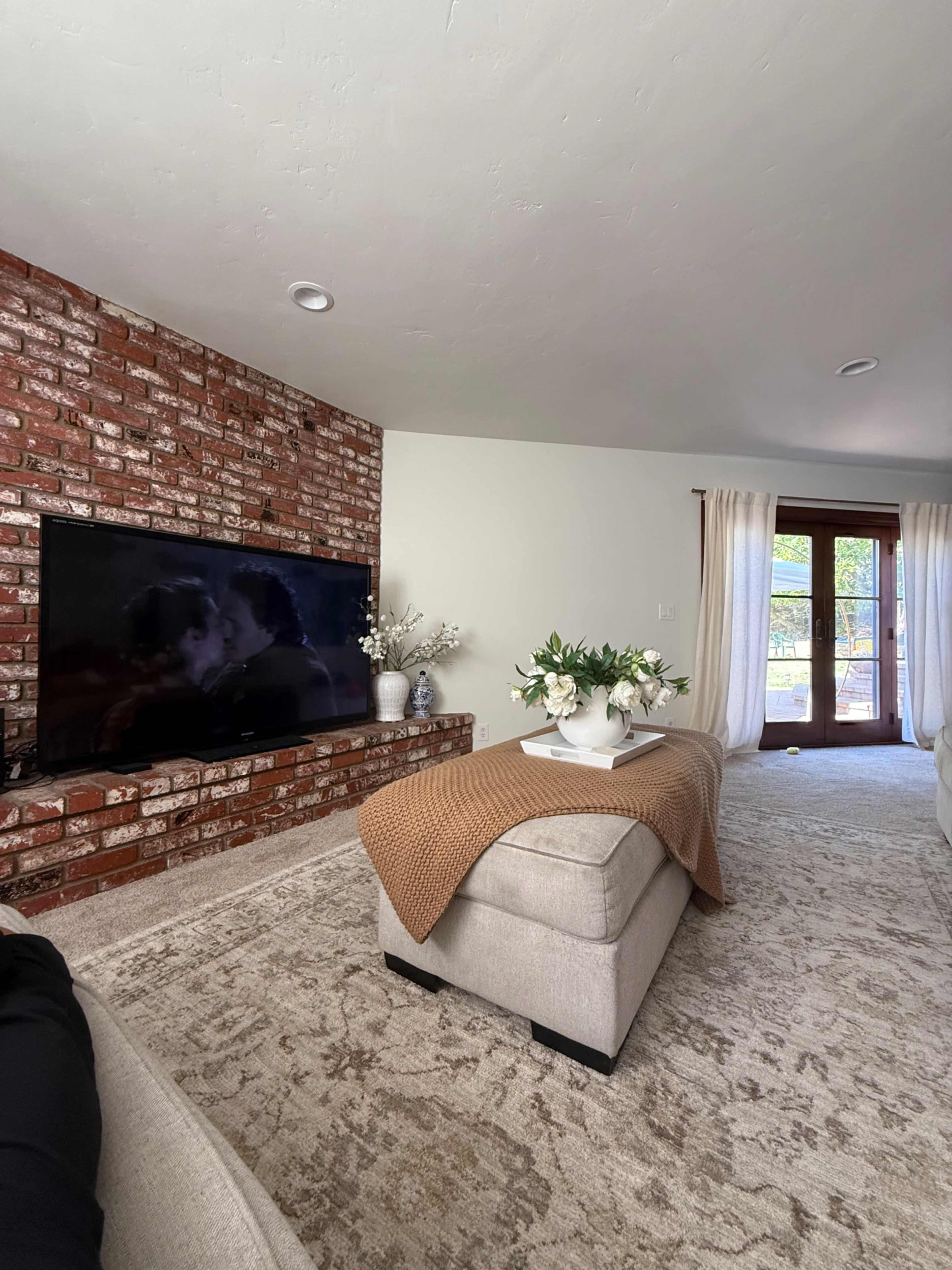 The living room features a brick accent wall, a large television mounted on a low platform, and a beige sectional sofa with a decorative blanket and a vase of flowers on a coffee table.